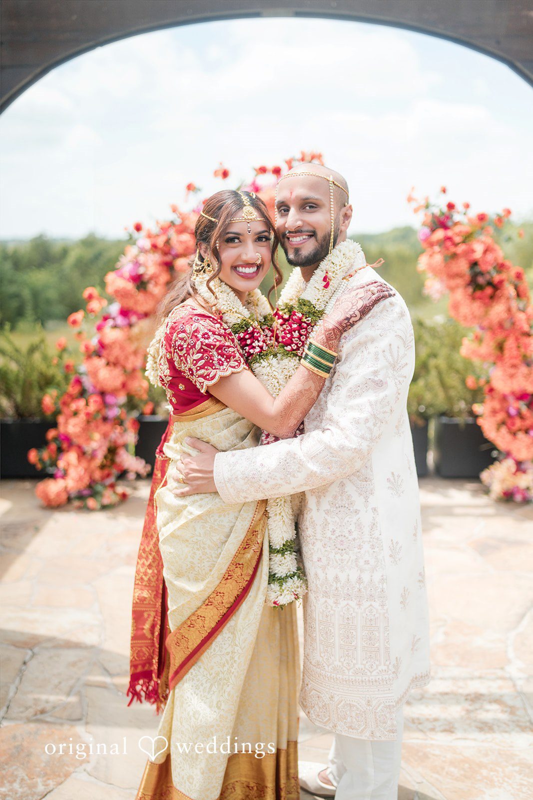 Groom holding the bride from behind while smiling in front of a red floral archway.
