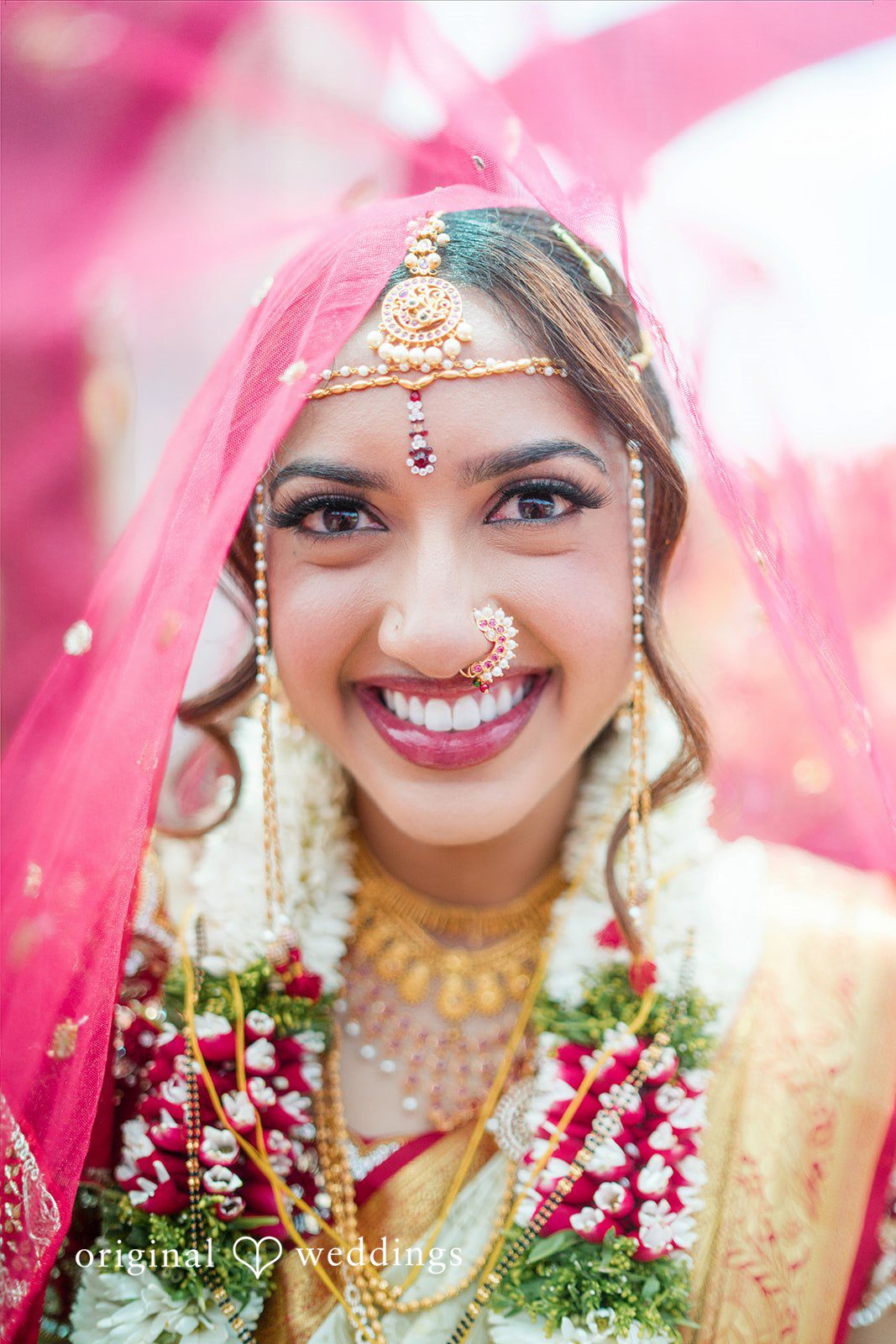A close-up portrait of a smiling bride wearing a red veil, traditional jewelry, and a floral garland.