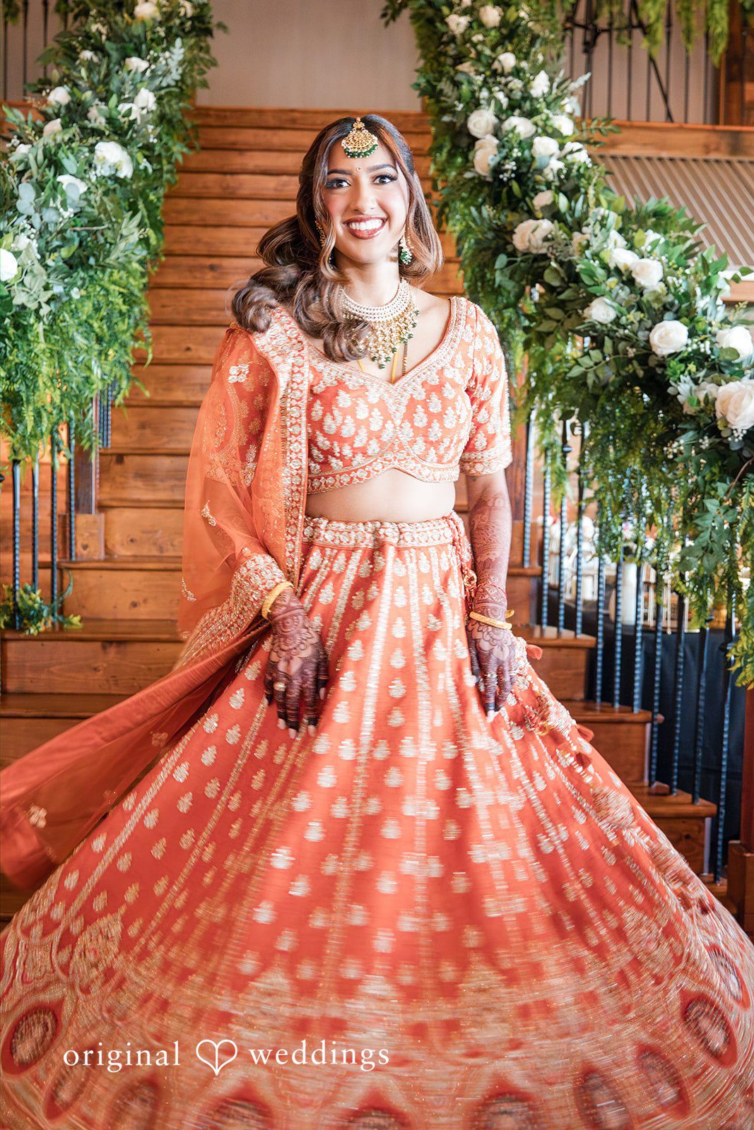 Bride wearing an orange lehenga standing on a decorated staircase decorated with greenery.