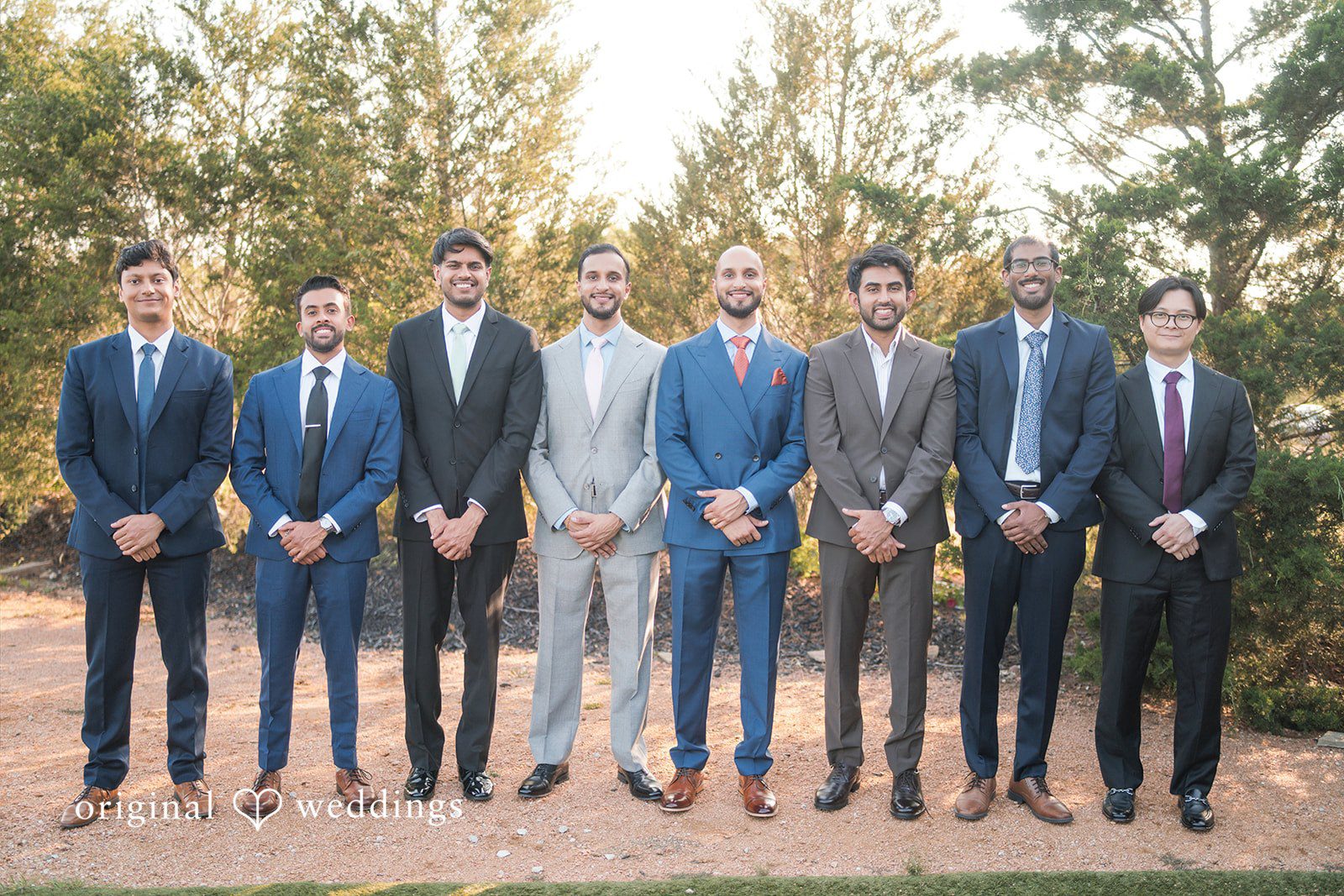 Groom and groomsmen dressed in various blue and grey suits posing together in a line outdoors.
