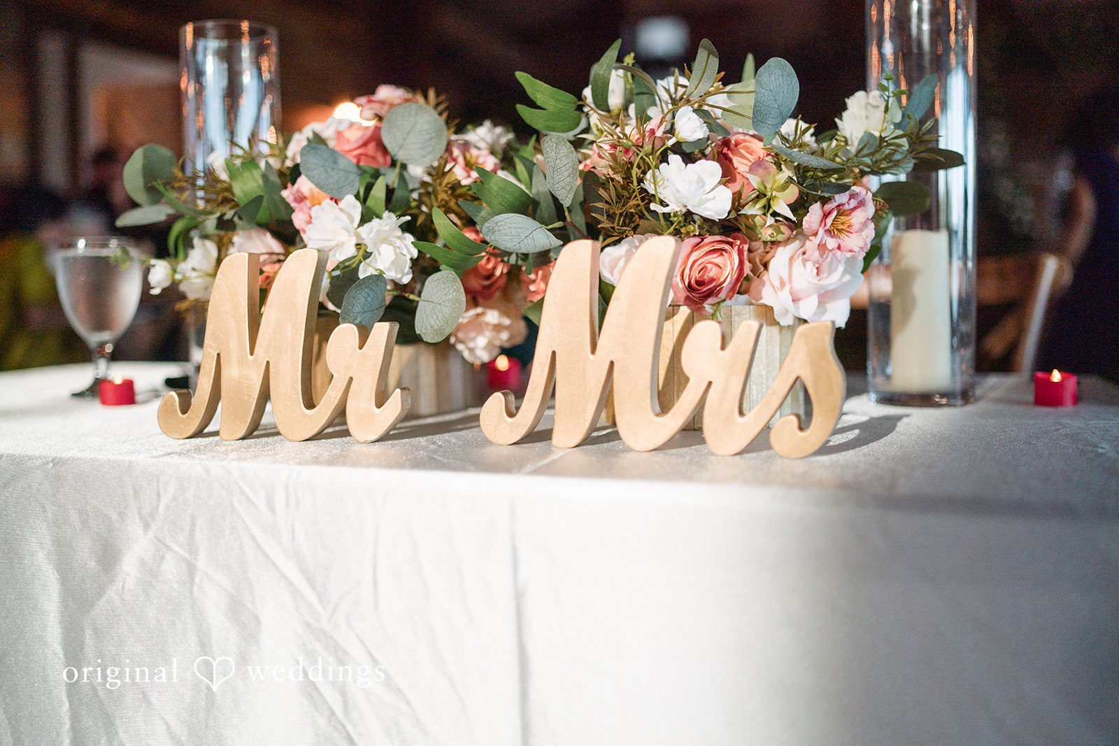 A wooden "Mr. & Mrs." sign placed on a white tablecloth with blurred floral decorations in the background.