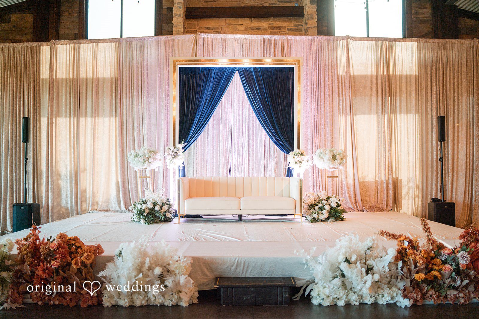 A wedding stage decorated with layered white drapes, a central blue fabric panel, and white floral arrangements.