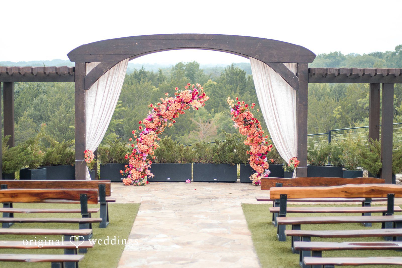 A wooden ceremonial arch decorated with a pink floral garland, standing before rows of wooden benches.