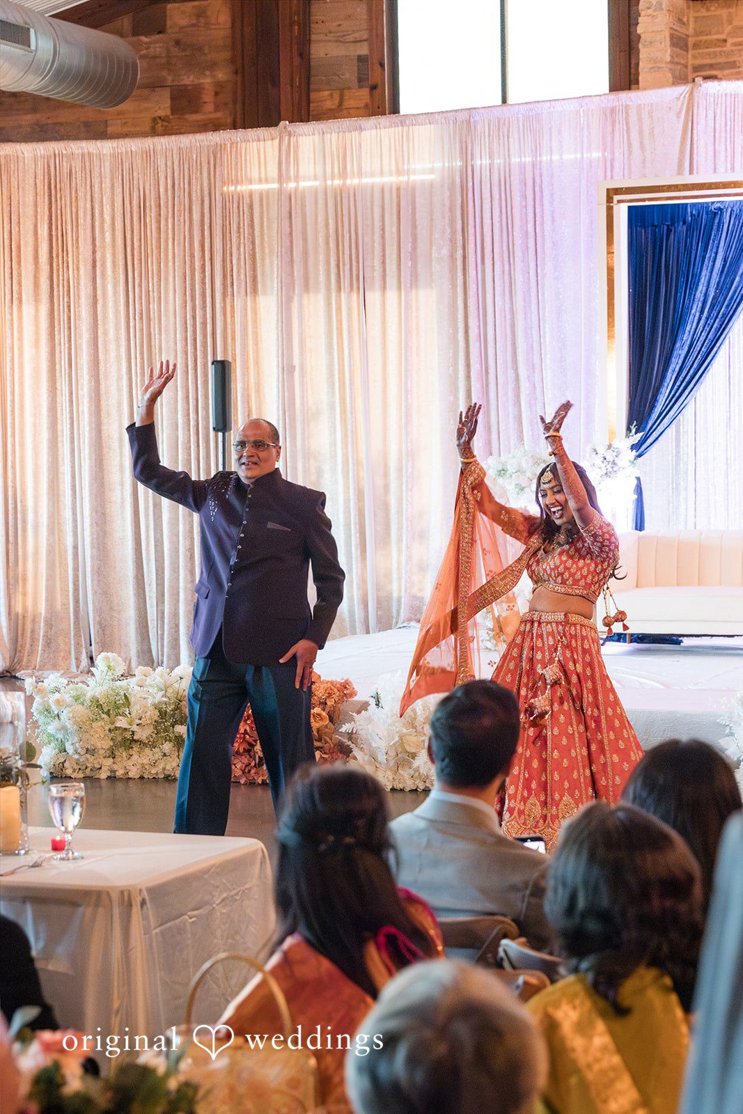 A man in a dark suit and a woman in an orange lehenga dancing on a stage in front of seated wedding guests.
