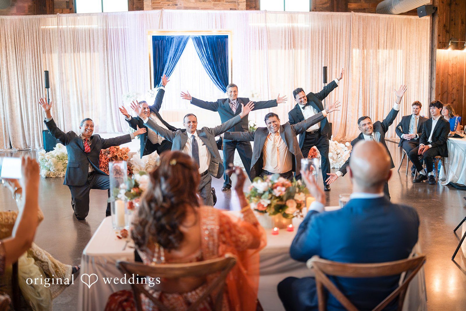 A group of men in suits strike a energetic dance pose with their arms wide open in front of a seated bride and groom.