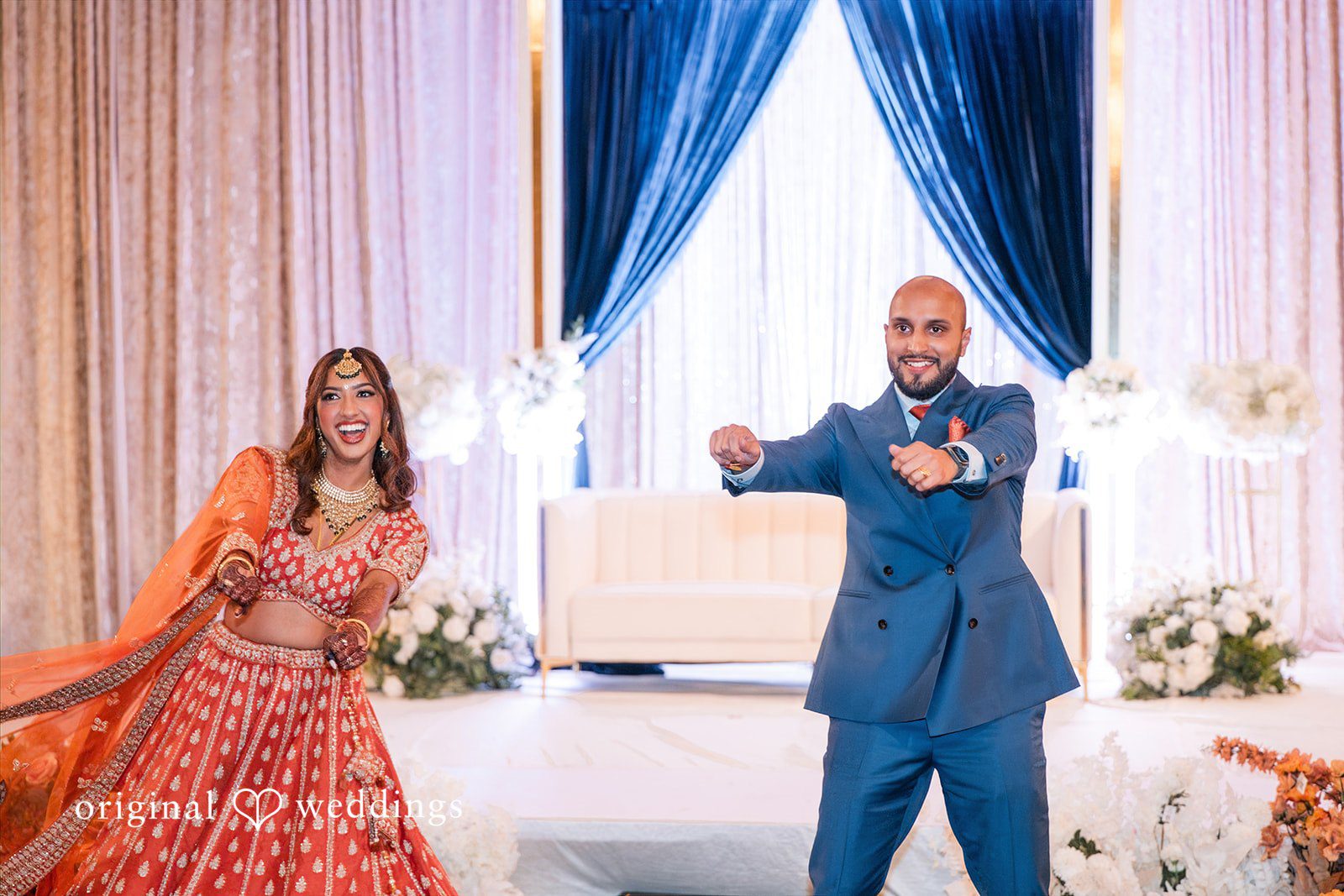 Bride in an orange lehenga and groom in a blue suit performing a dance on a stage.