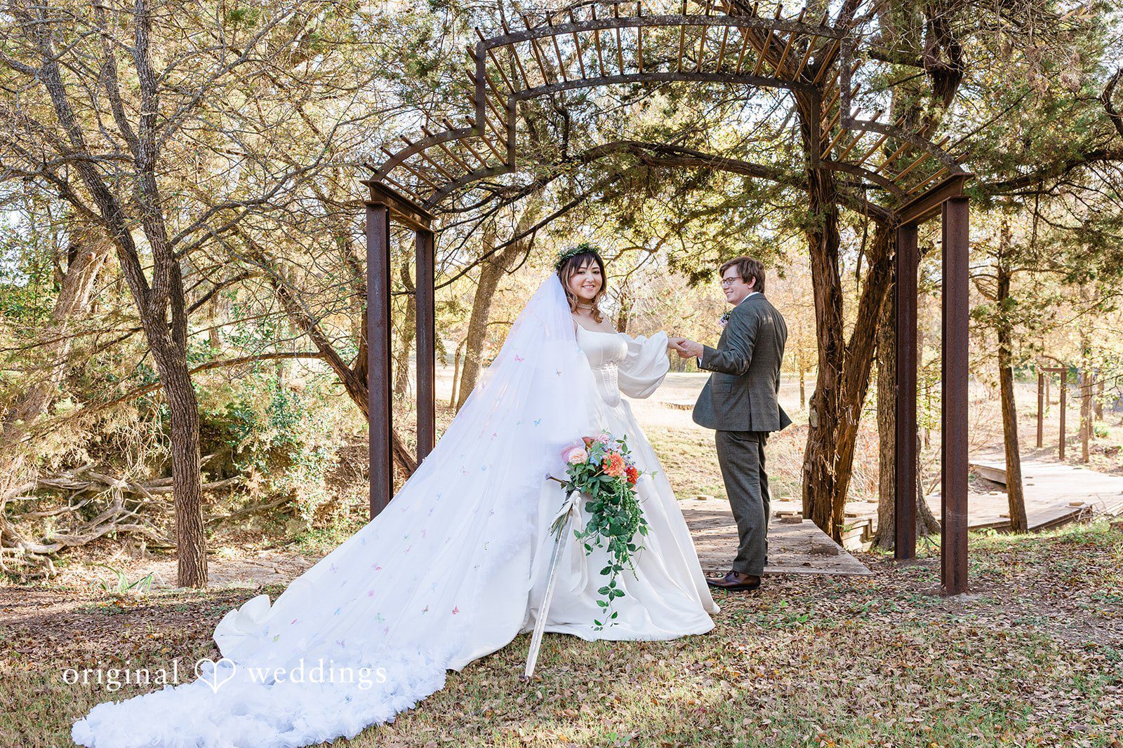 November 15, 2025 – Brittney & Benjamin- (ID-3986) A bride in a long, flowing white veil and gown standing with her groom in a wooded area.