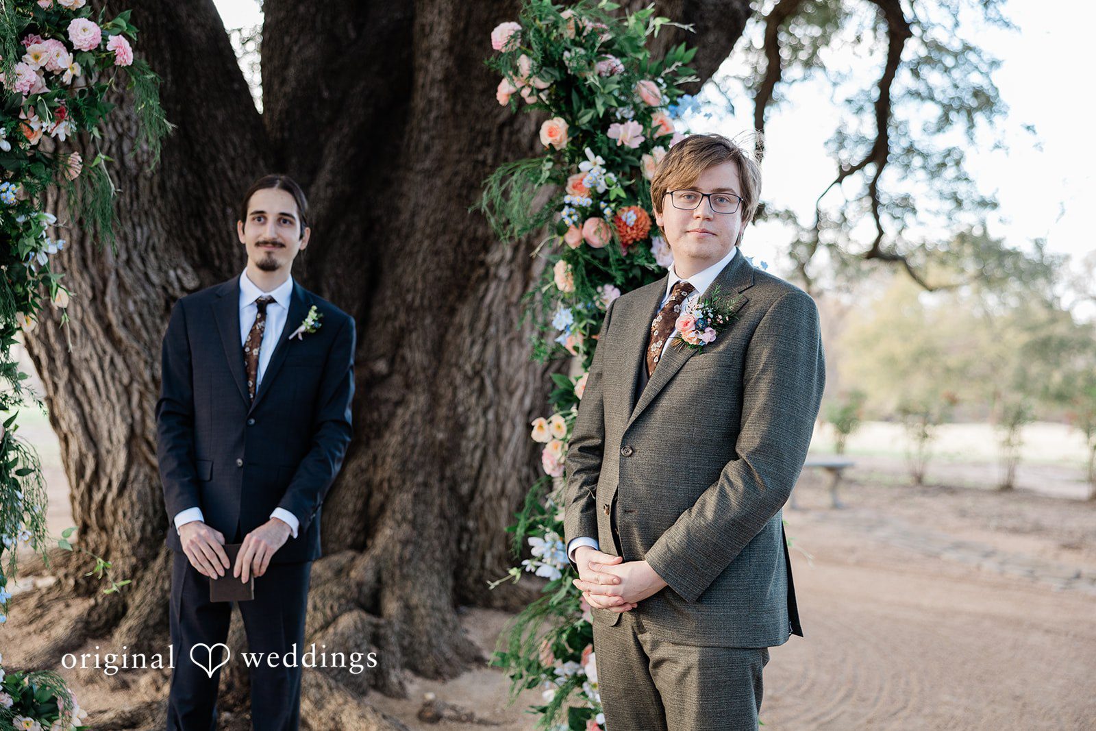 Brittney + Benjamin A groom in a grey suit stands with the wedding officiant in a dark suit in front of a floral arch.