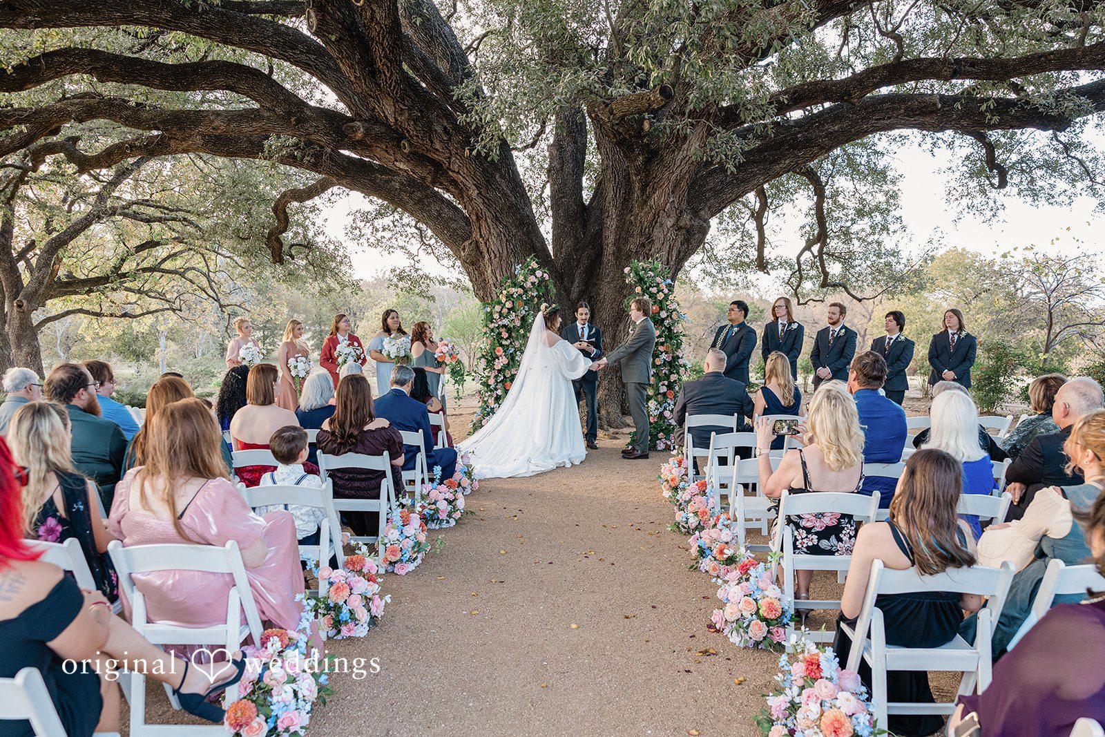 stonebridge-wedding-event-venue-airy-dallas-brittney-benjamin-286-7w4a3446-12 A wedding ceremony taking place under a massive, wide-spreading tree with seated guests.