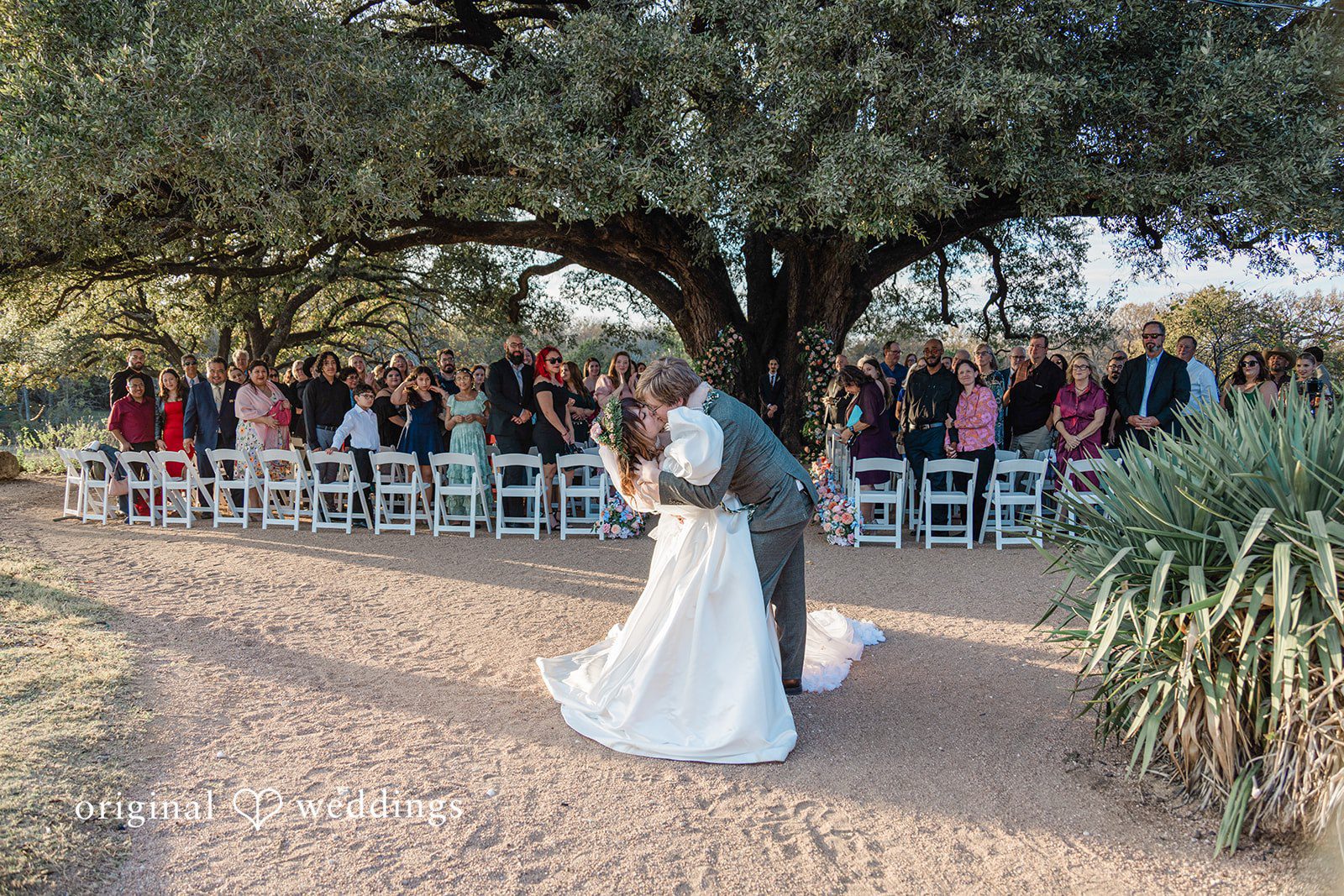 Brittney + Benjamin A bride and groom sharing a kiss on a dirt path in front of seated guests.