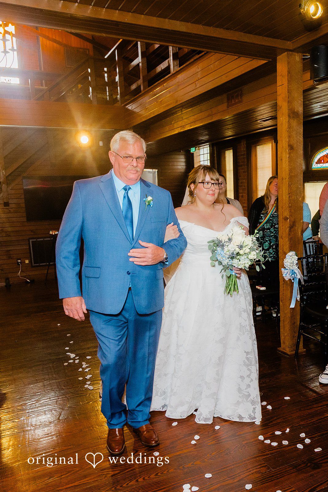 A bride in a white wedding dress being walked down the aisle by a man in a blue suit.