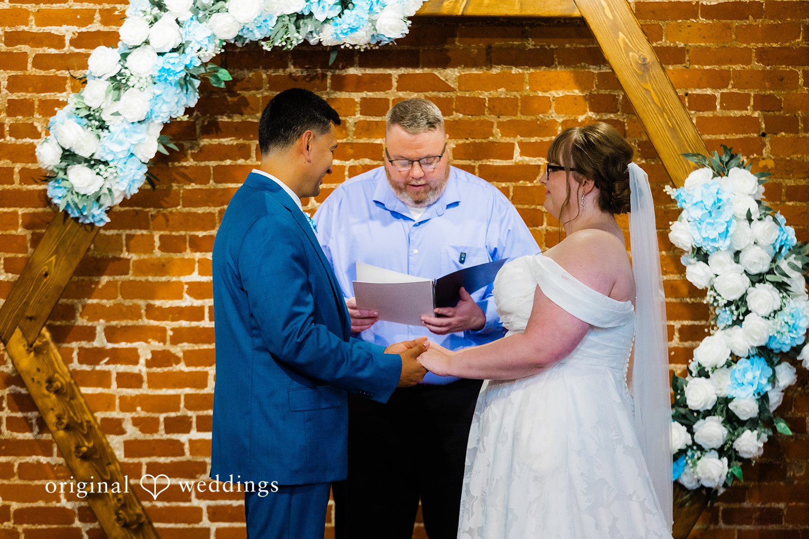 A bride and groom holding hands during their wedding ceremony while an officiant reads.