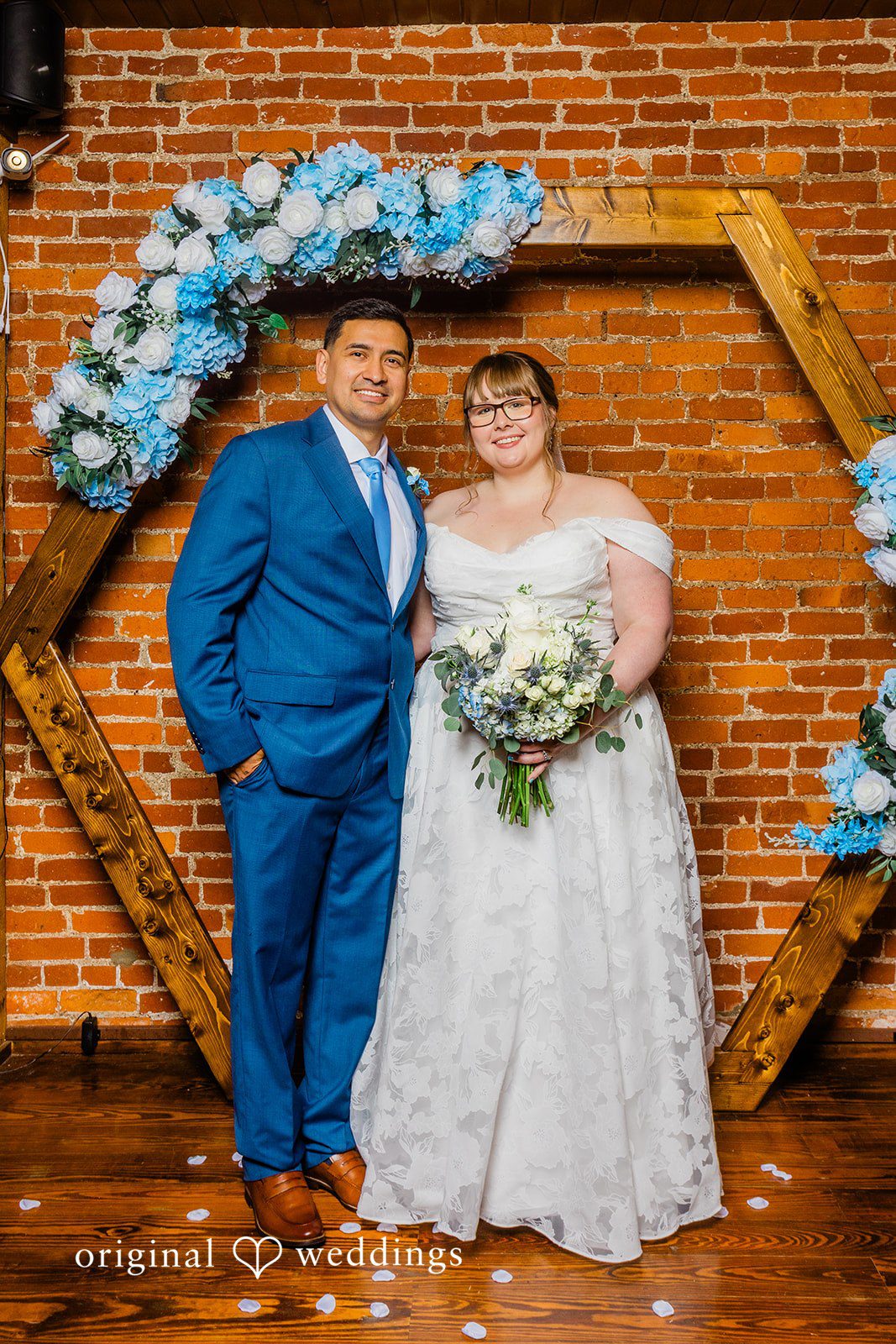 A bride and groom posing together in front of a  wooden arch with blue flowers.
