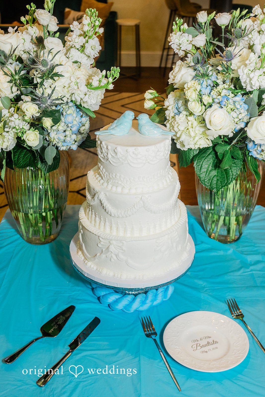 A three-tiered white wedding cake displayed on a blue tablecloth alongside floral arrangements.