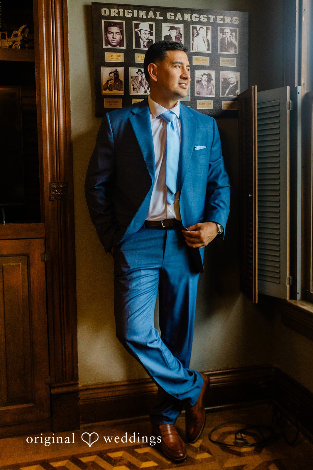 A groom wearing a bright blue suit leaning against a dark wooden wall.