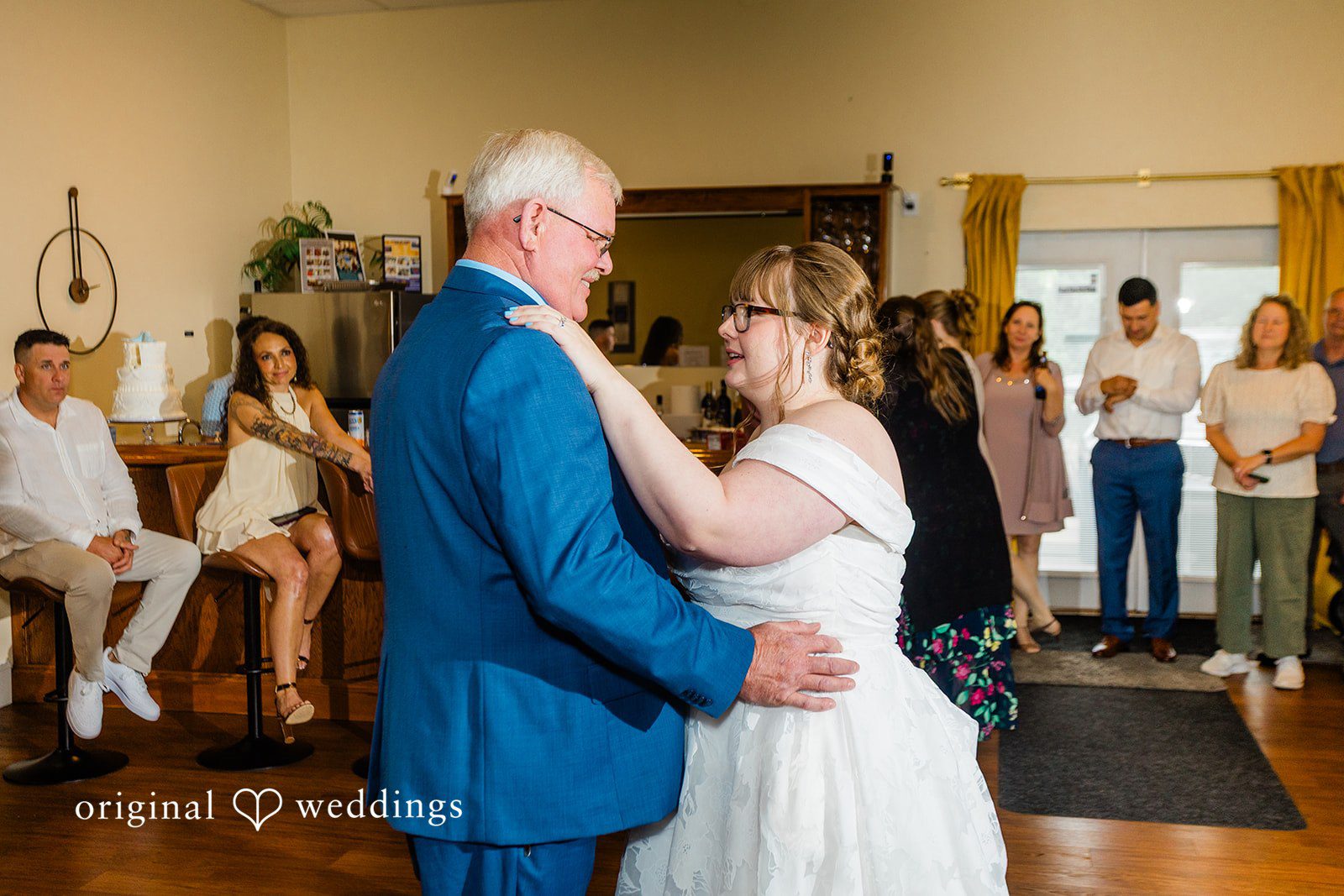 A bride dancing with a man in a blue suit while guests watch in the background.