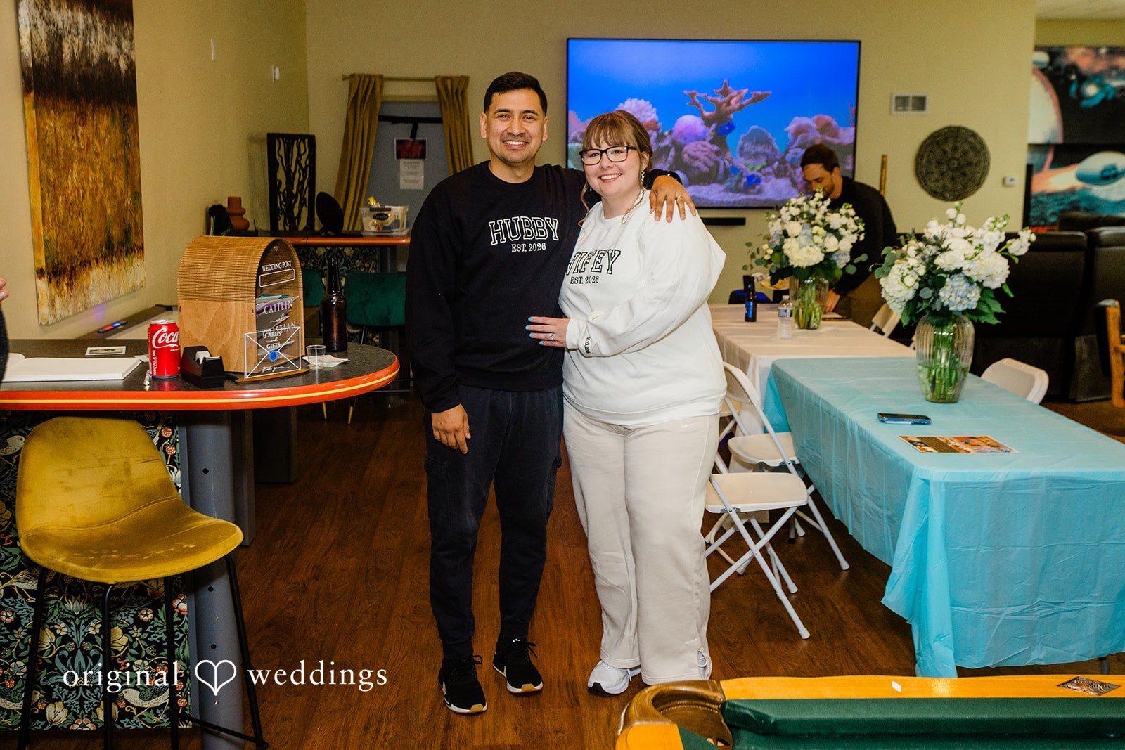 A smiling man in a black "HUBBY" sweatshirt and a smiling woman in a white "WIFEY" sweatshirt pose together at a reception.