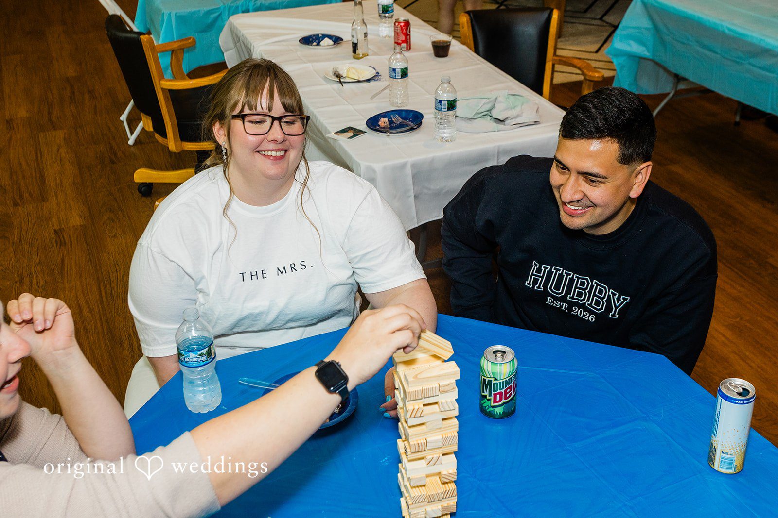 A bride and groom sitting at a blue table playing a block-stacking game.
