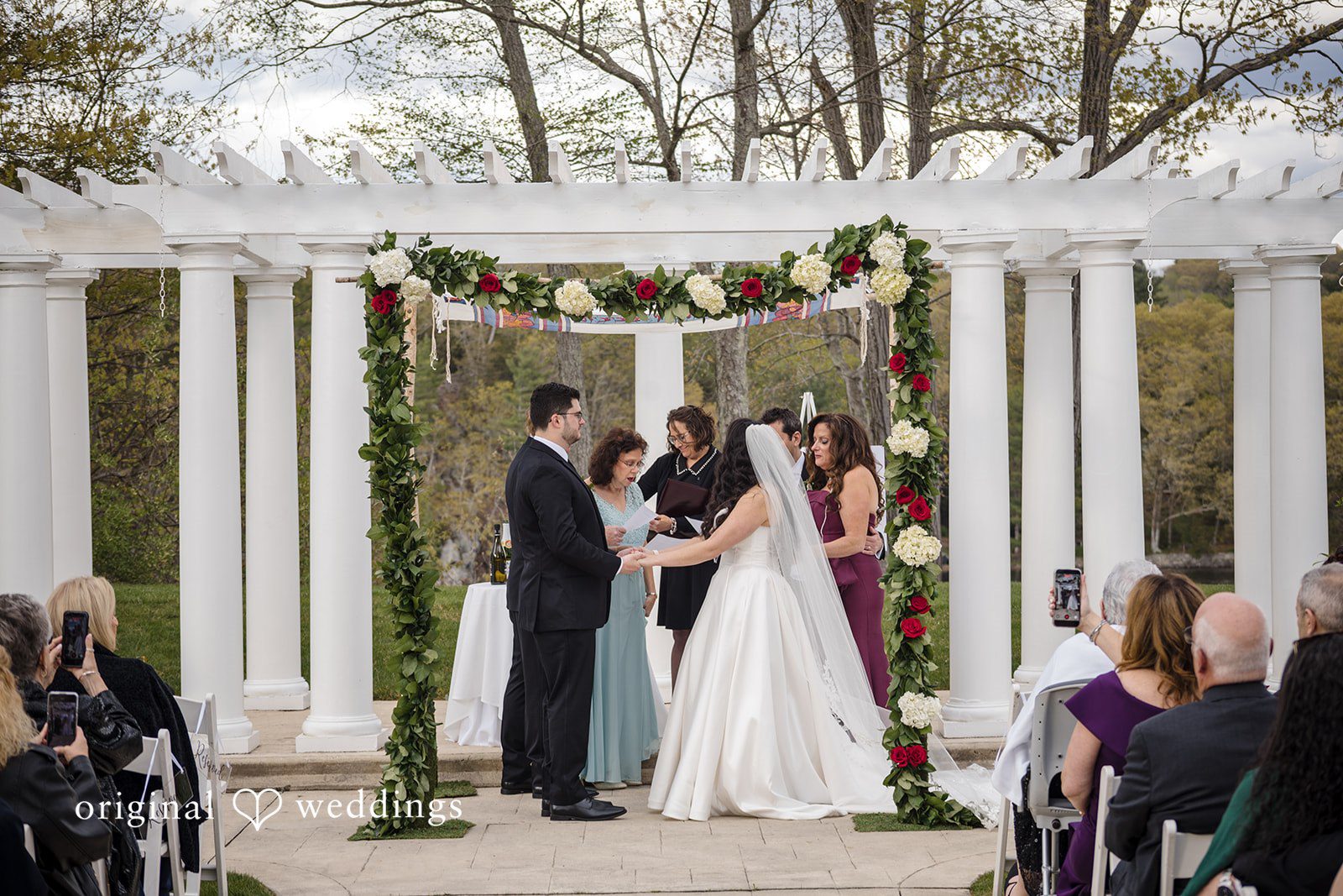 Bride and groom hold hands while exchanging vows beneath floral chuppah.