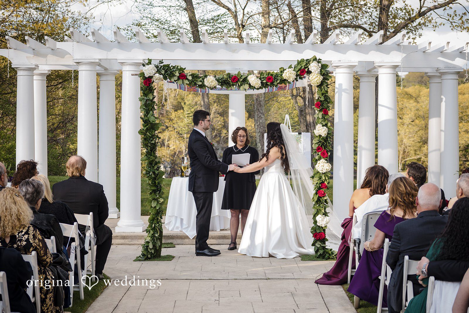 Bride and groom exchange vows under floral arch surrounded by guests.