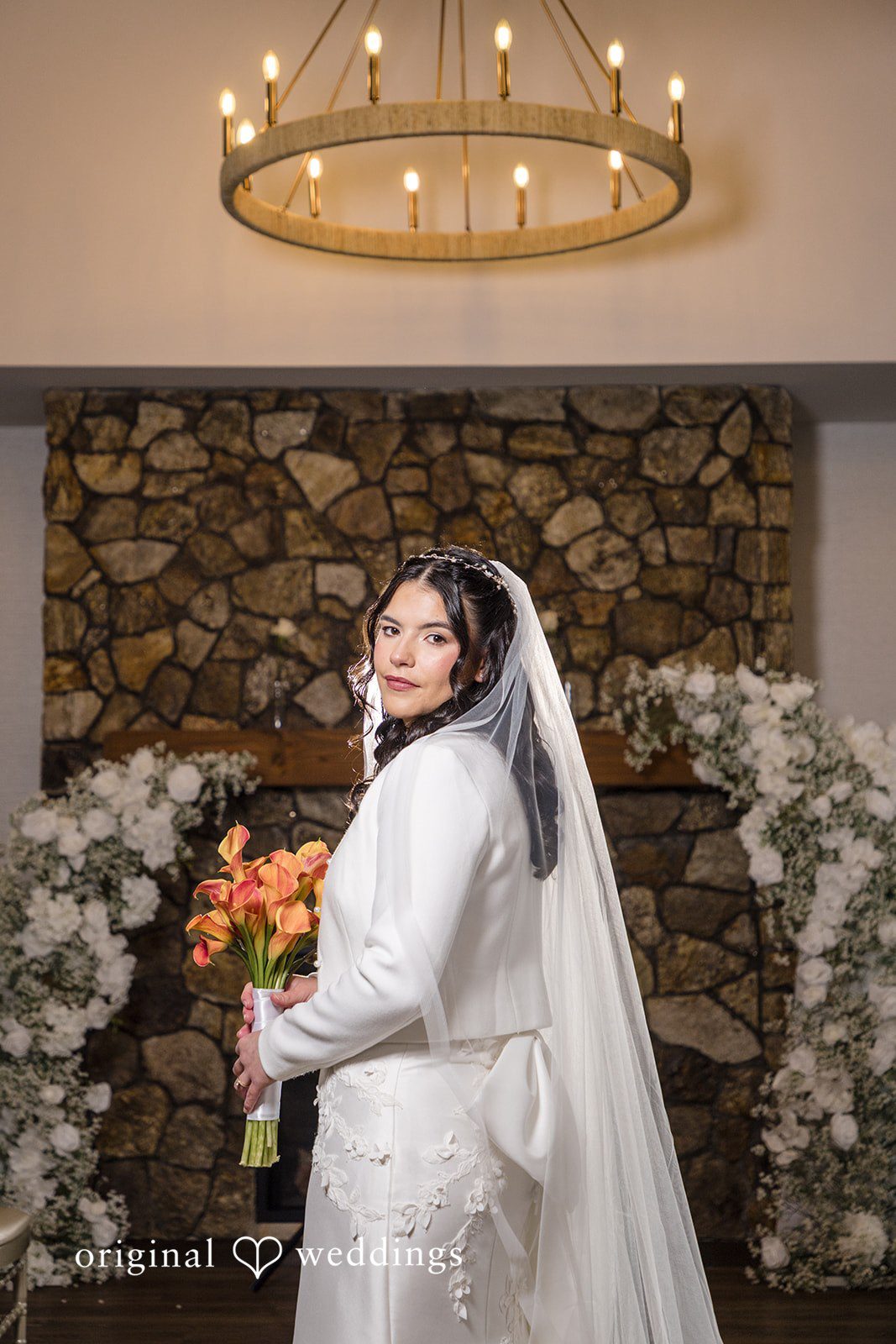 Maria + Eddy A side profile view of a bride holding an orange bouquet near a stone fireplace.