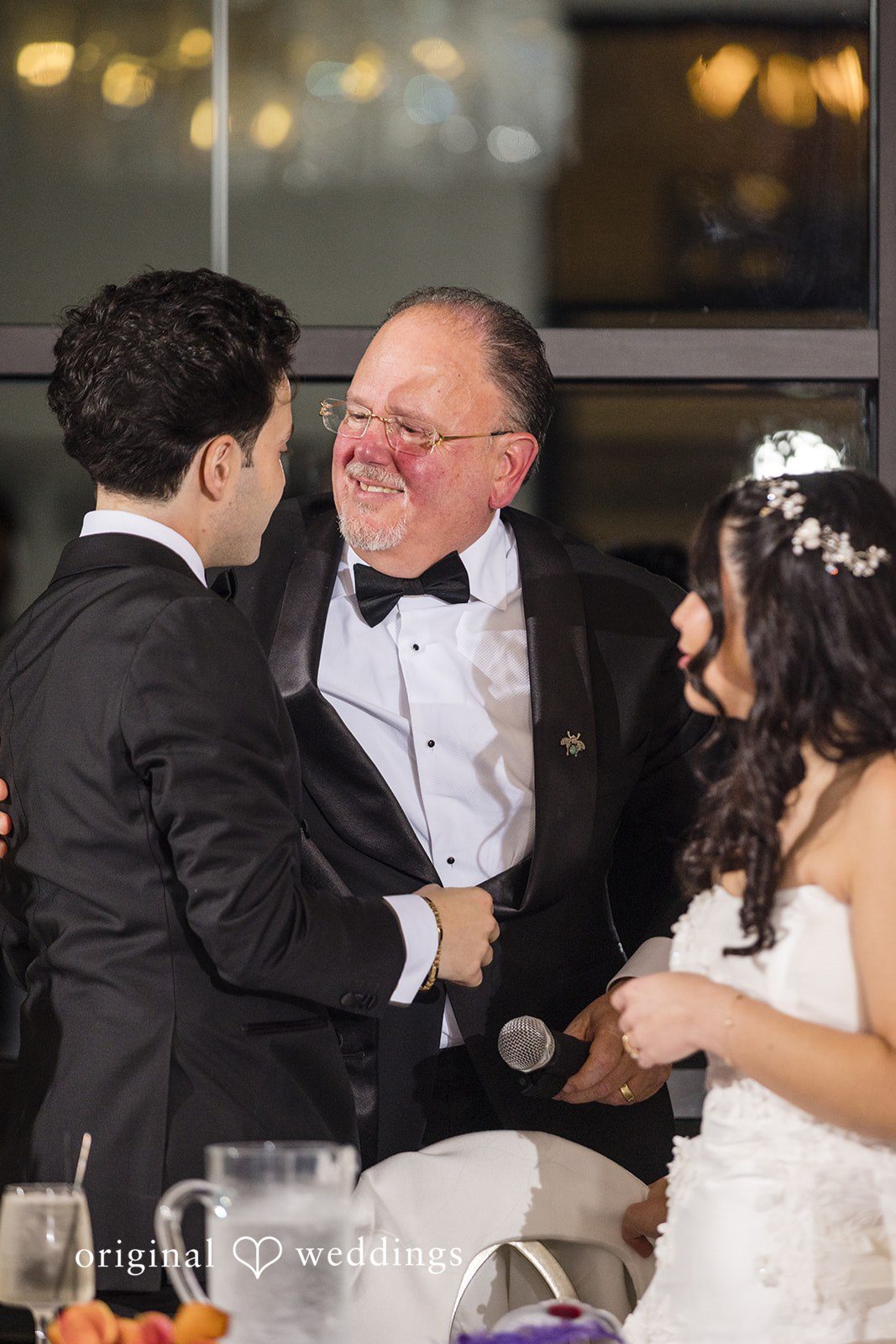 Maria + Eddy A smiling man in a tuxedo holding a microphone embraces a groom while a bride stands nearby.