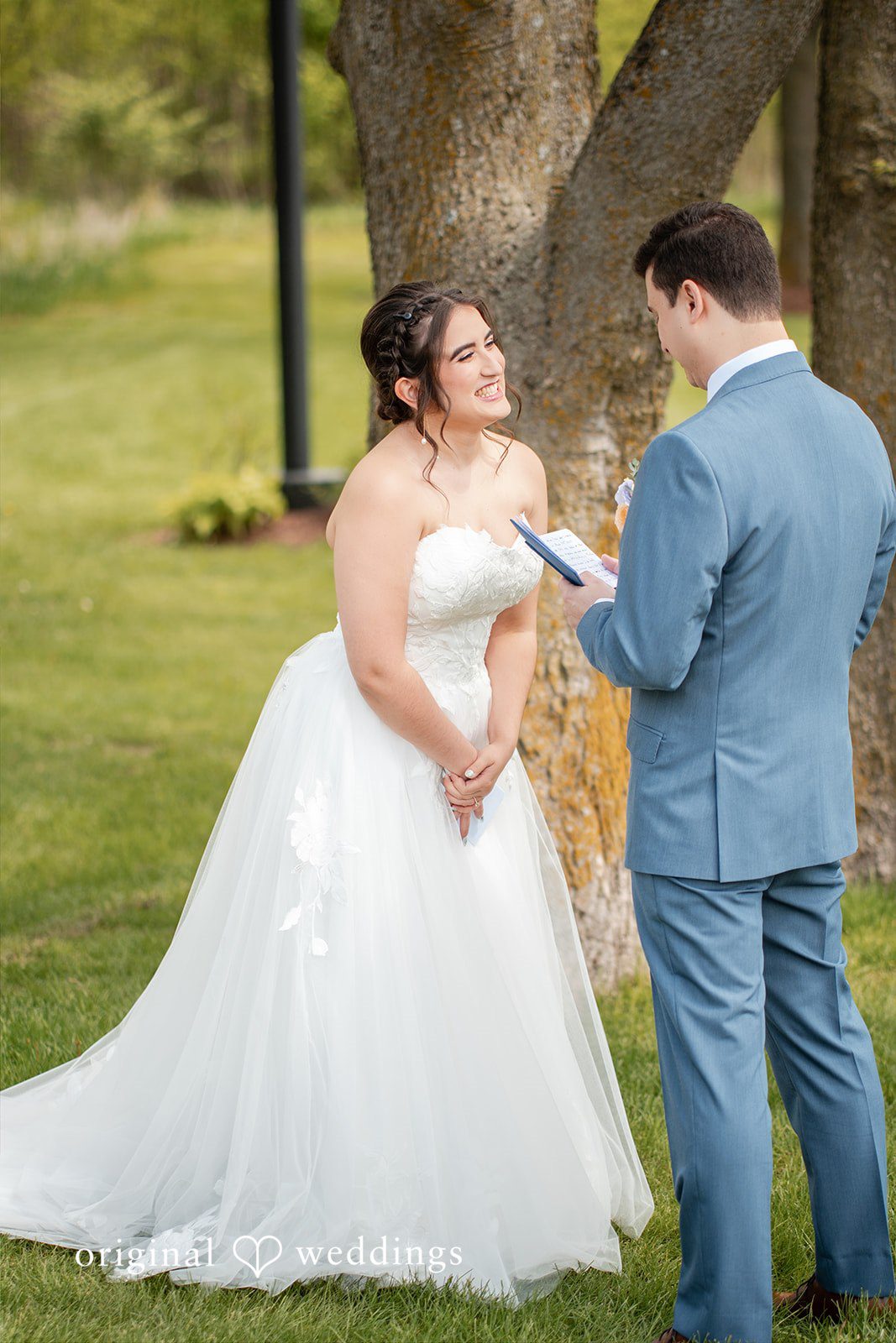 Bride smiling as groom reads vows during an outdoor wedding ceremony.