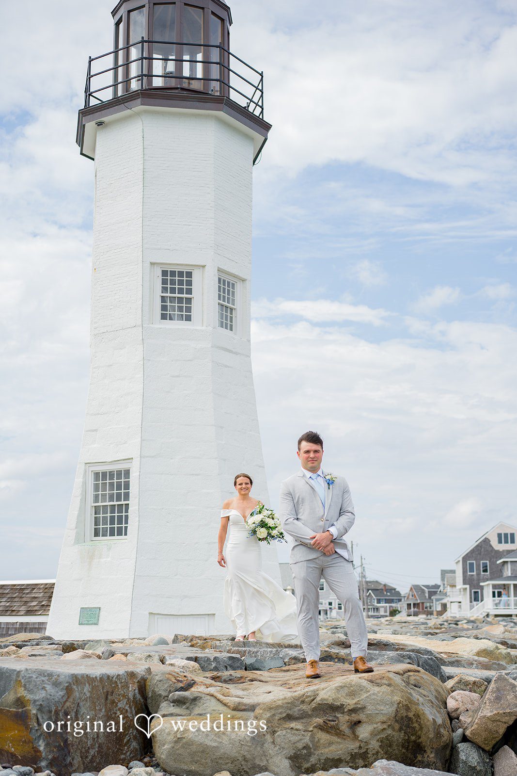 Molly + Joe A bride and groom stand together on rocks near the ocean with a lighthouse behind them.