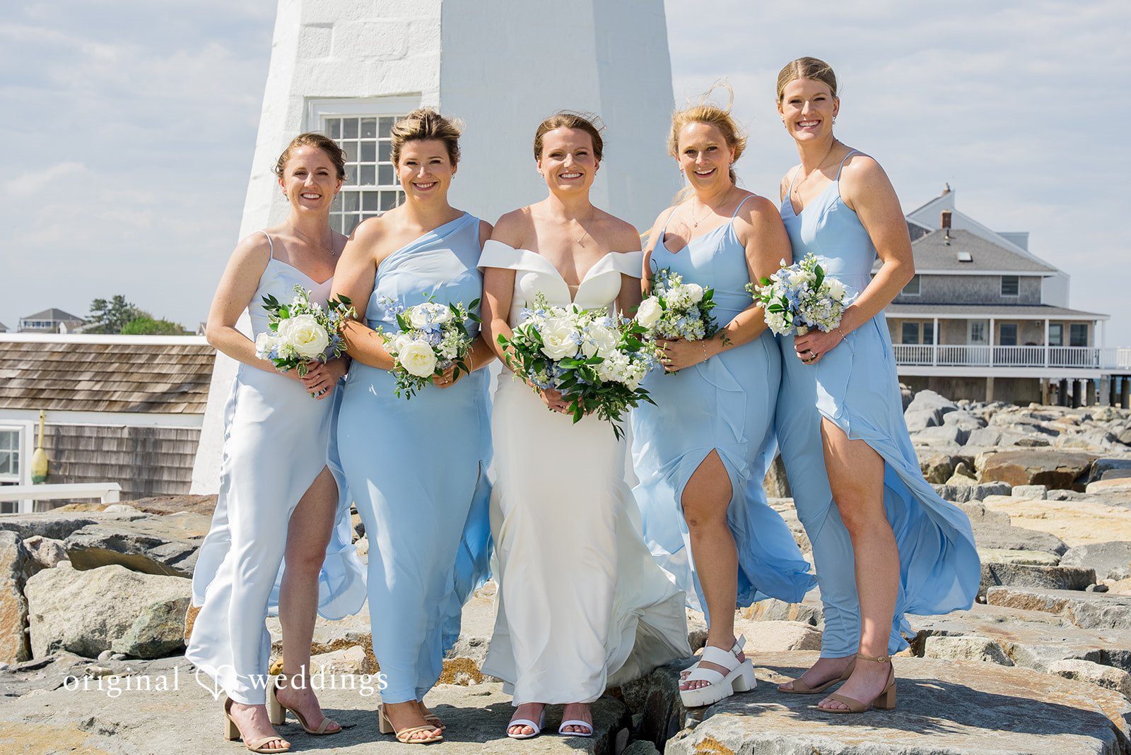 Molly + Joe A bride and bridesmaids wearing light blue dresses pose together near a rocky coast.