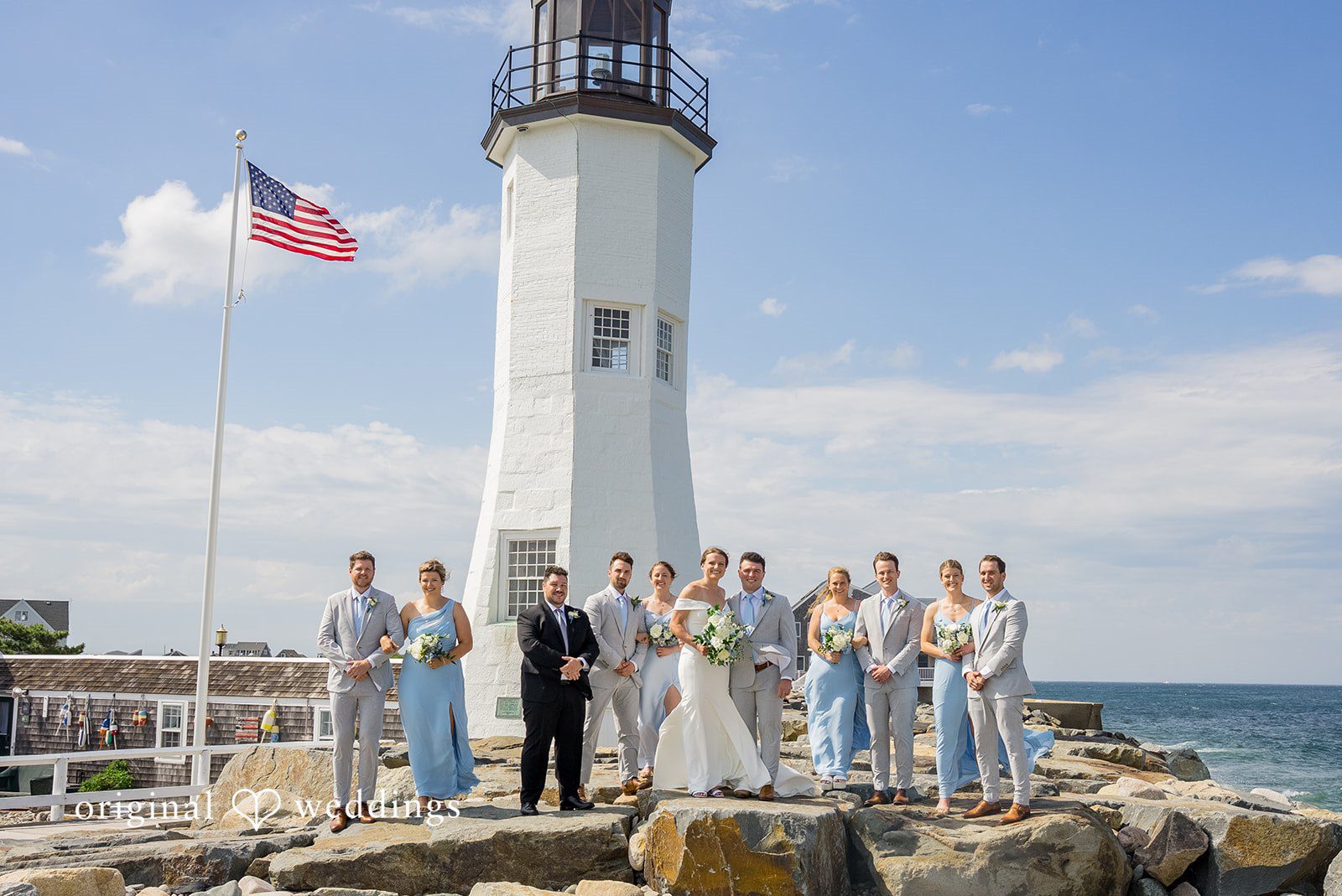 Molly + Joe A wedding party poses together at the base of a tall white lighthouse.