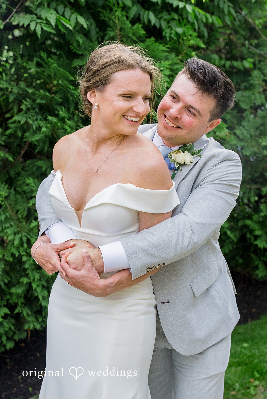 Molly + Joe A groom embraces the bride from behind as they smile together in a garden setting.