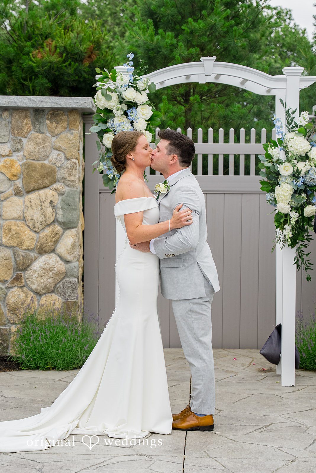 Molly + Joe A bride and groom embrace and share a kiss by a stone wall gate.