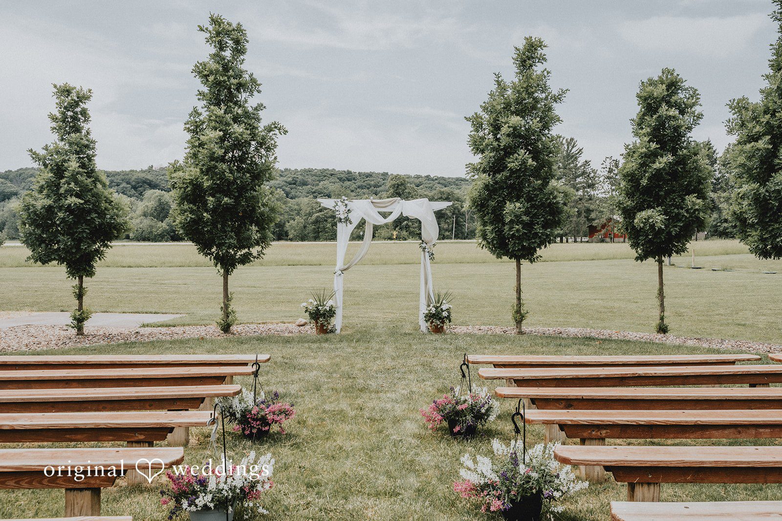 Neil + Hallie A wedding aisle lined with benches leading to a simple archway on a lawn.