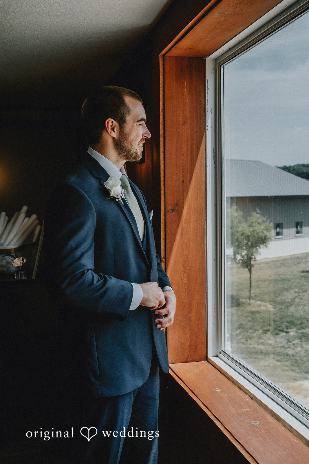 Neil + Hallie A groom standing by a large window looking out toward the wedding venue.