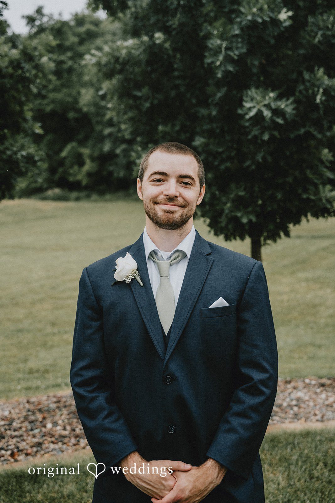 Neil + Hallie A groom in a navy blue suit smiling while posing on a grassy lawn.