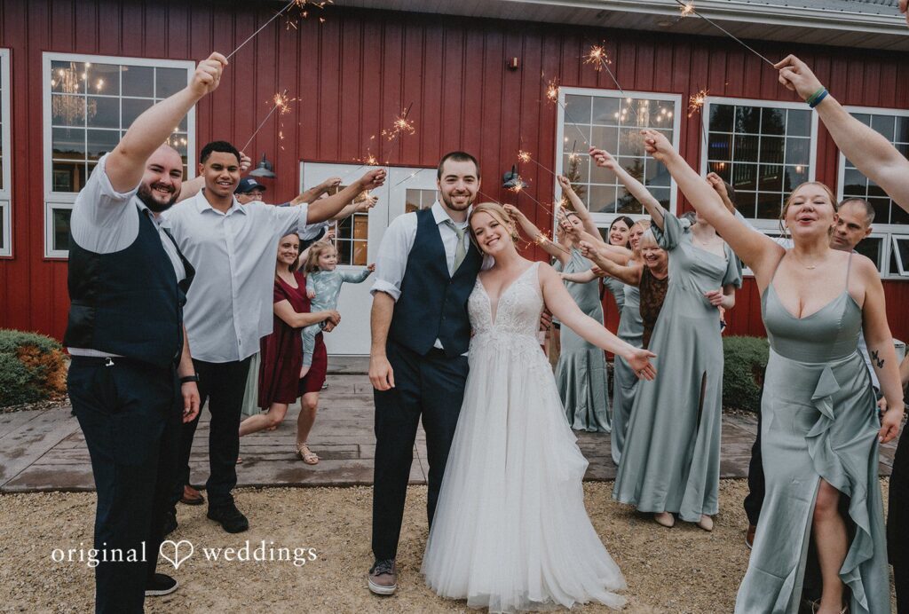 Neil + Hallie A newlywed couple walking through a cheering crowd of wedding guests holding up light sticks at Bearwood Event Barn captured by Minneapolis Wedding Photographers from Original Weddings