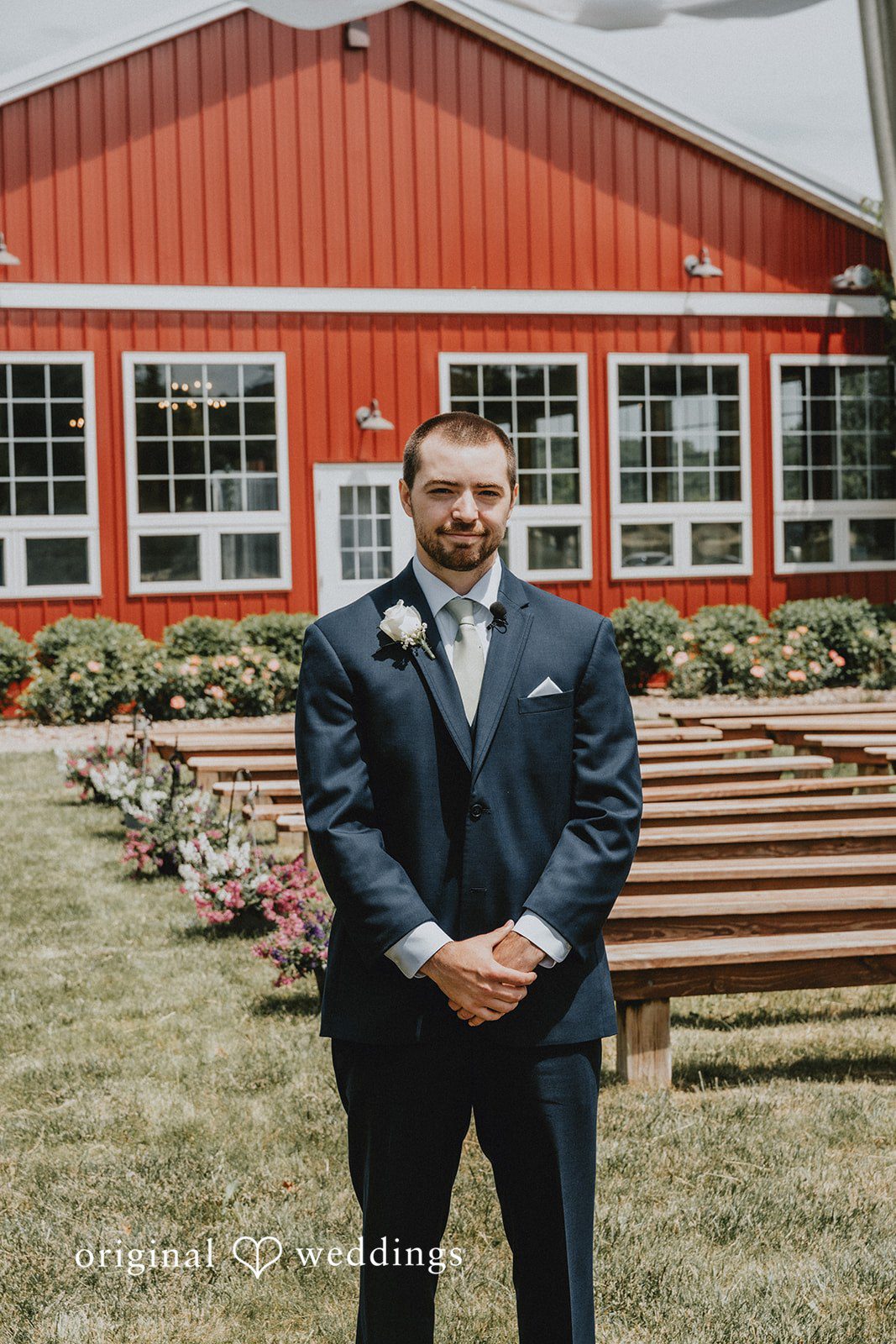 Neil + Hallie A groom in a suit standing on wood stairs in front of a red building.