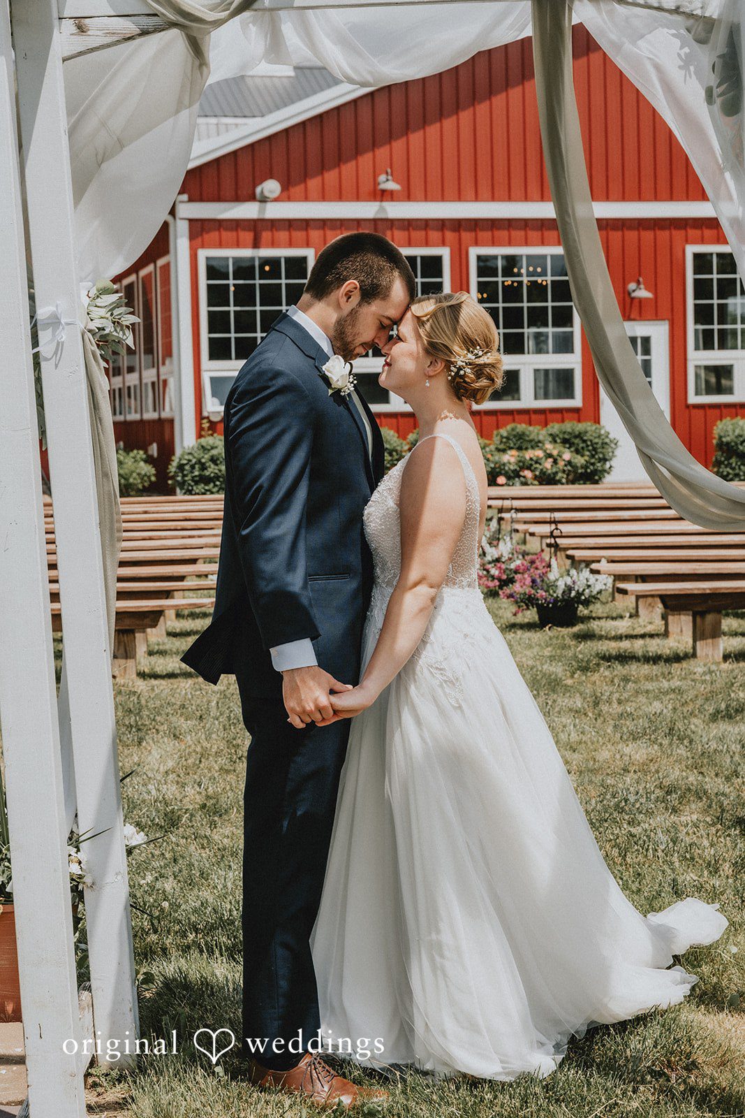 Neil + Hallie A bride and groom embracing and smiling at each other in front of a red barn structure.
