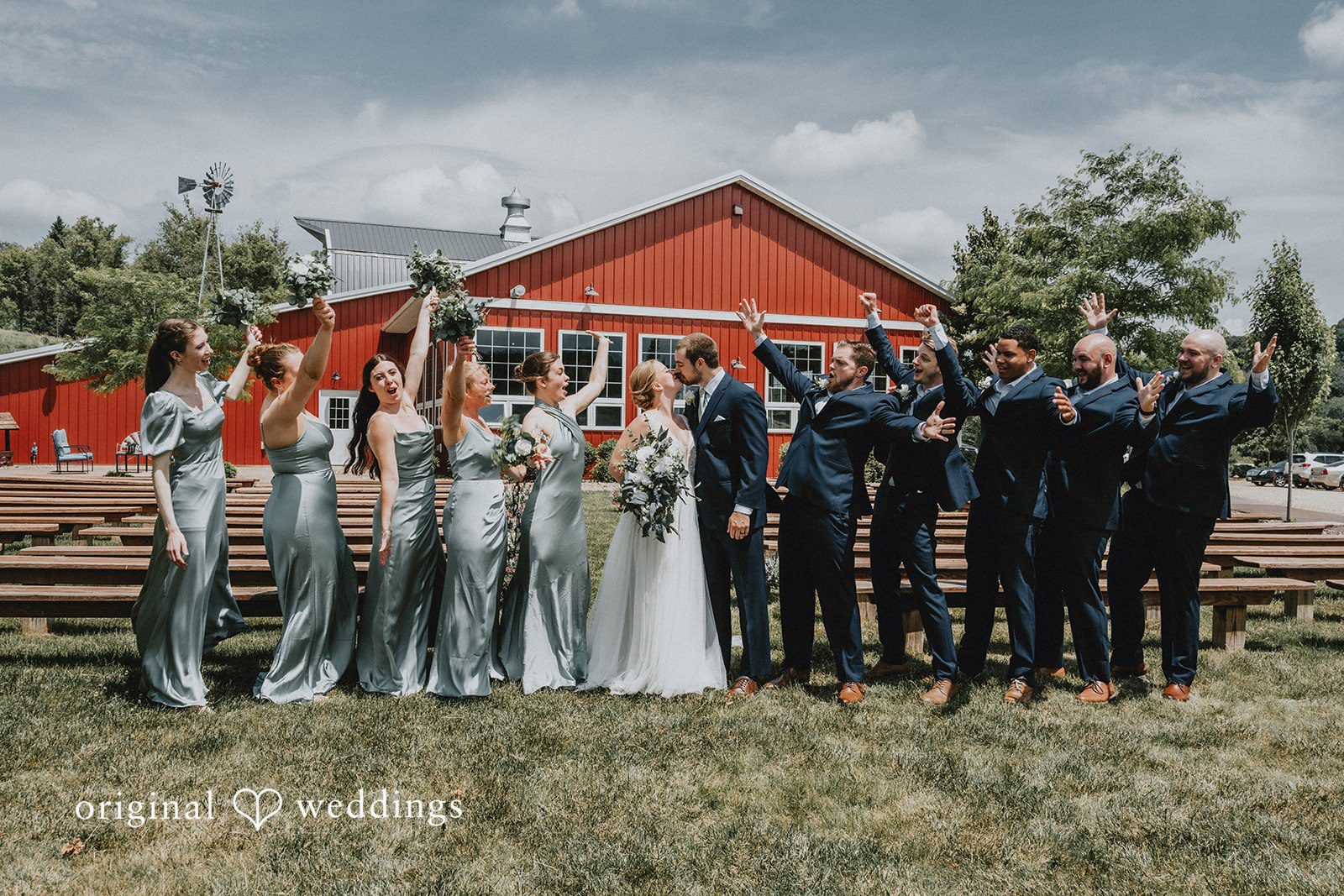 Neil + Hallie The full wedding party posing in a long line on a lawn with a red barn in the background.