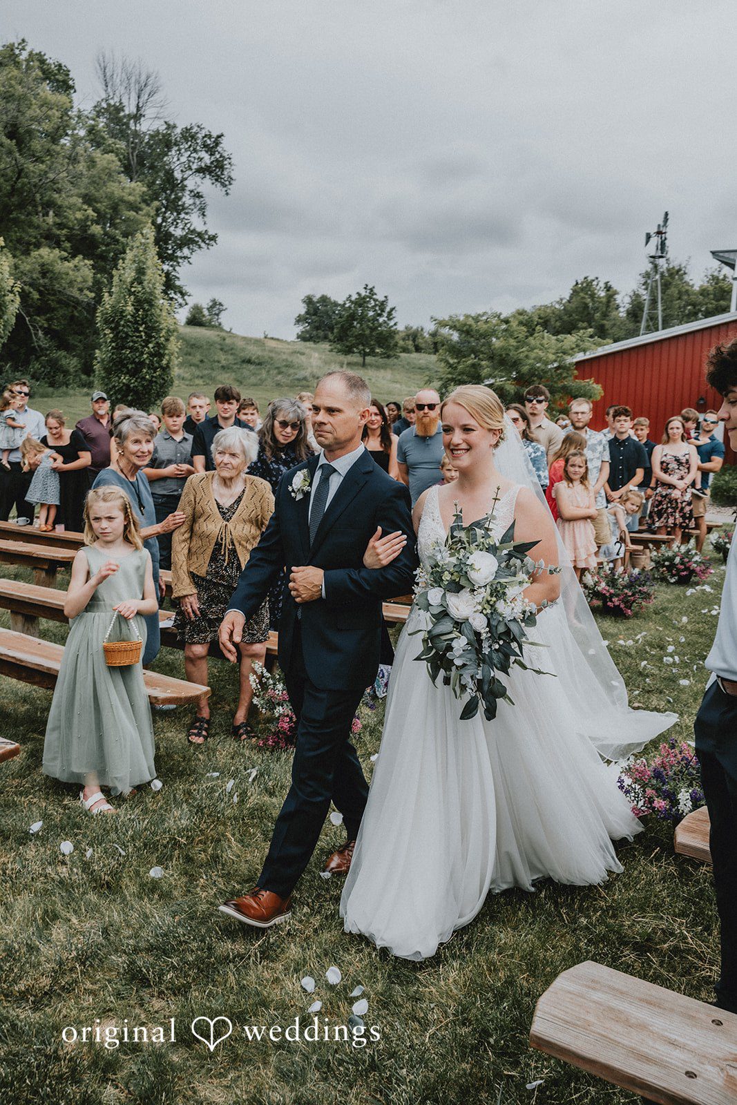 Neil + Hallie A bride walking down a grassy wedding aisle lined with seated guests.