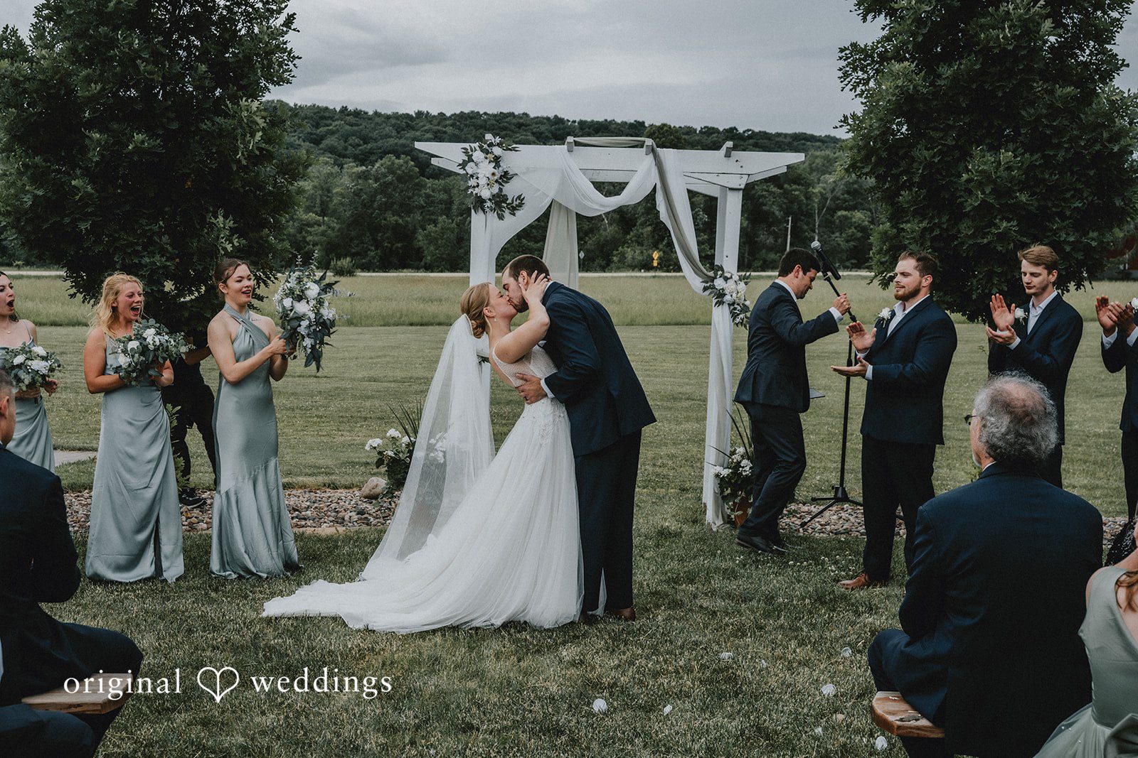 Neil + Hallie Couple sharing first kiss and the full wedding party posing in two rows on a grassy lawn.