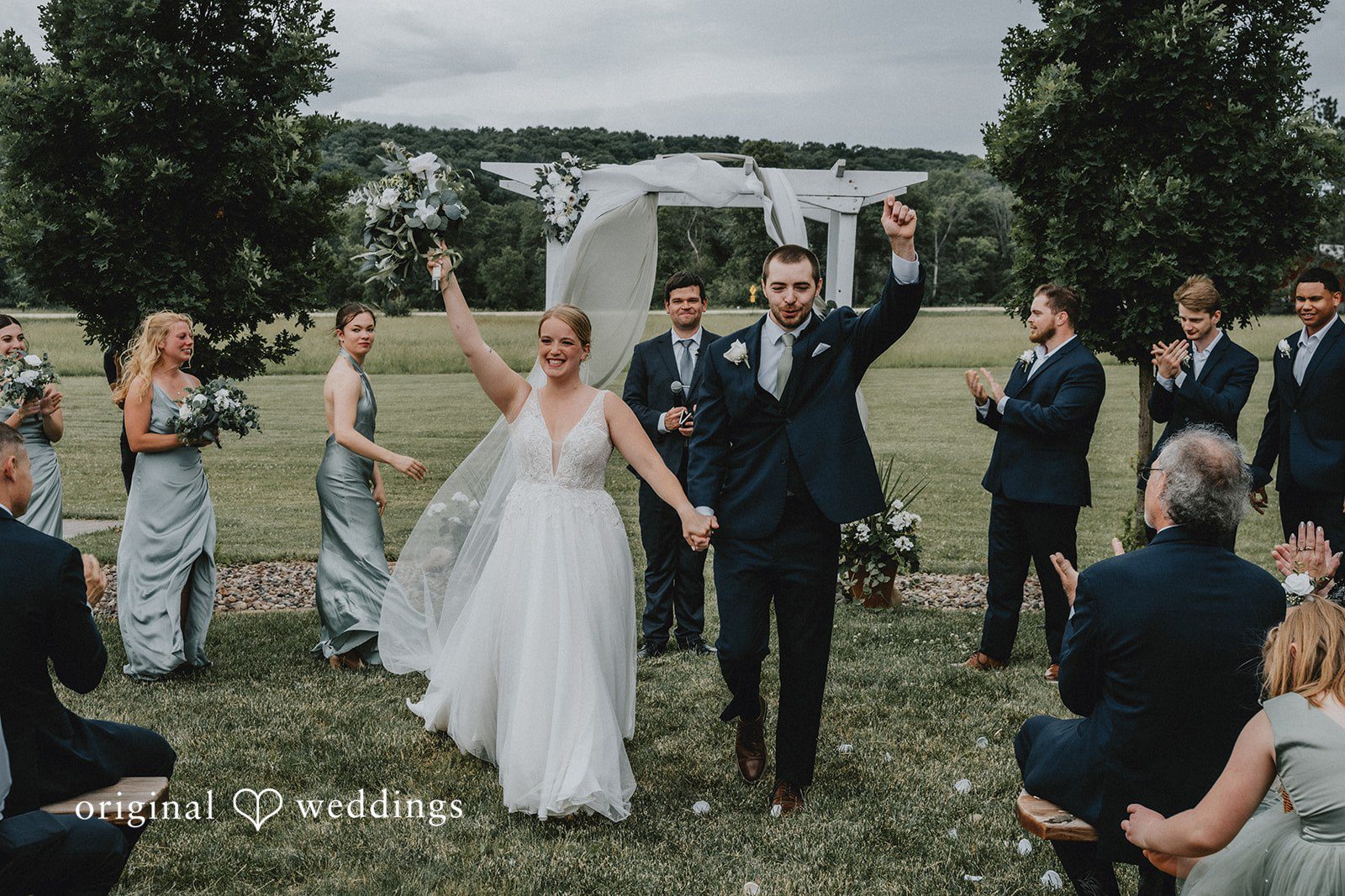 Neil + Hallie A newlywed couple celebrating with raised arms while walking down the aisle.
