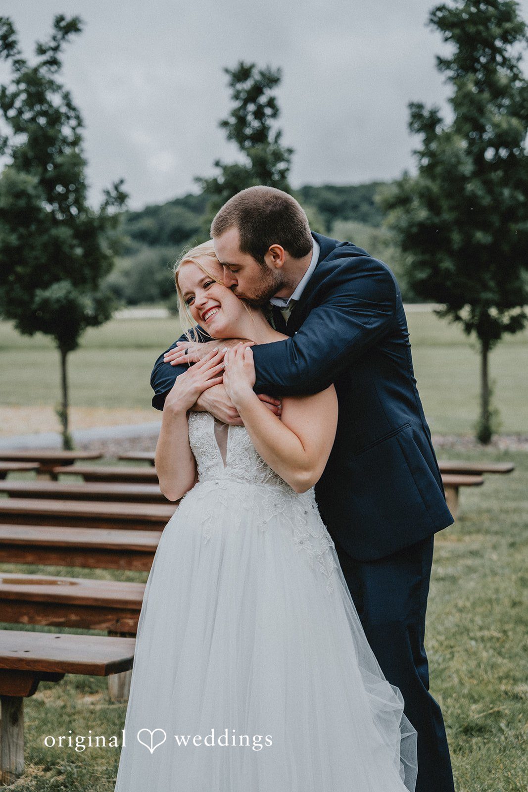 Neil + Hallie A groom affectionately embracing his smiling bride on a grassy field.