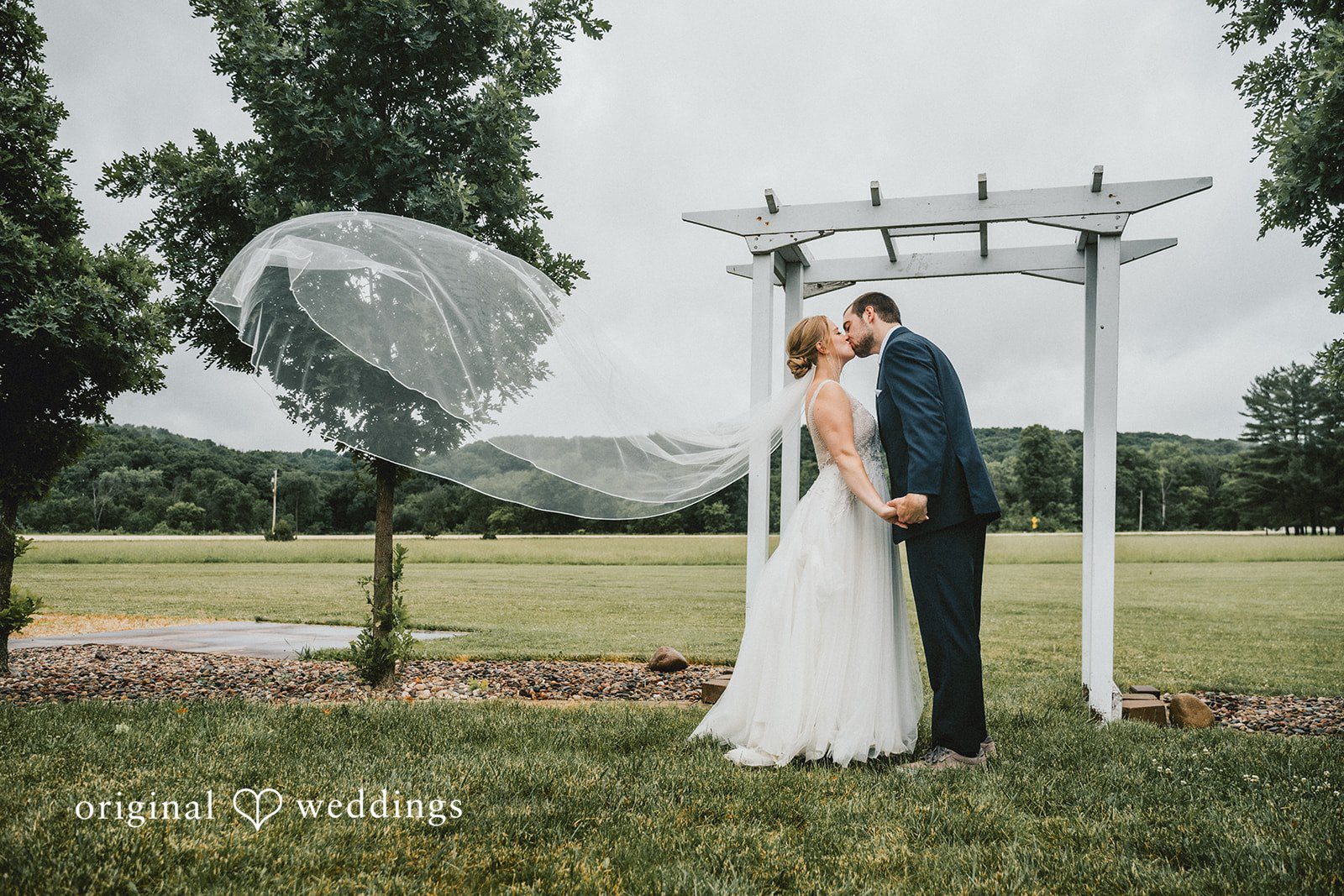 Neil + Hallie A bride and groom sharing a kiss under a white arch structure on a lawn while her veil catches the wind.