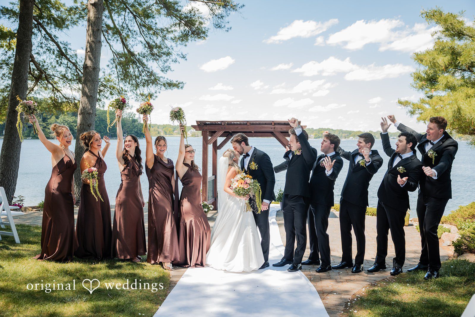 Trey + Bailey Outdoor wedding party picture featuring the bride and groom sharing a kiss with friends. at Blake’s Backyard captured by Detroit Wedding Photographers from Original Weddings.