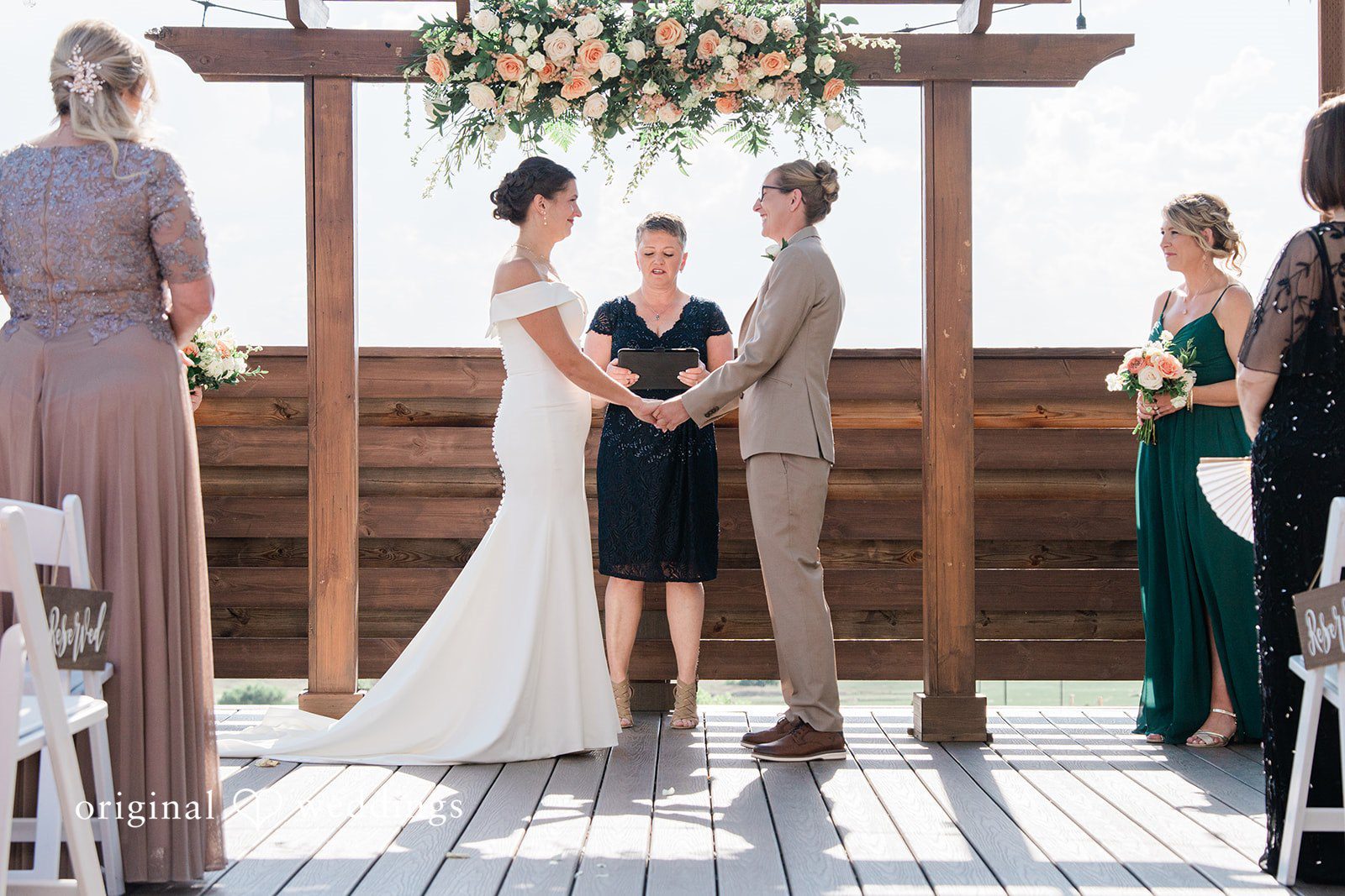 Rachel & Krystal A wedding ceremony taking place under a wooden pergola with guests looking on.