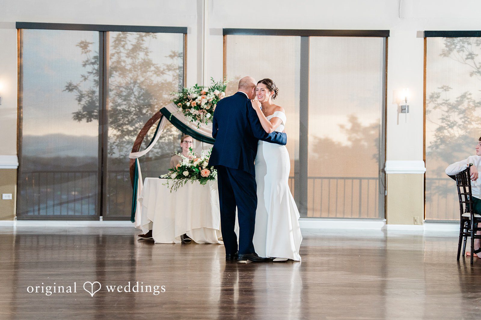 Rachel & Krystal A bride in a white wedding gown dancing with a man in a navy blue suit on a polished wooden dance floor during a reception.
