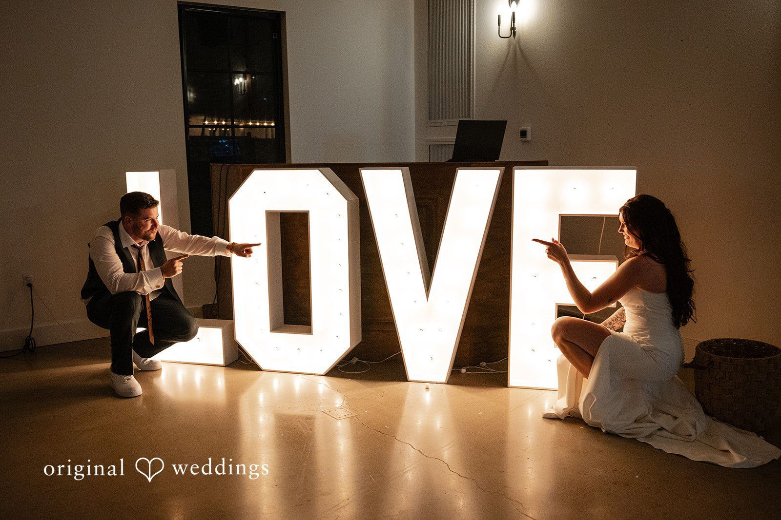 Noah + Jade A bride and groom posing playfully next to large, glowing marquee letters that spell "LOVE".