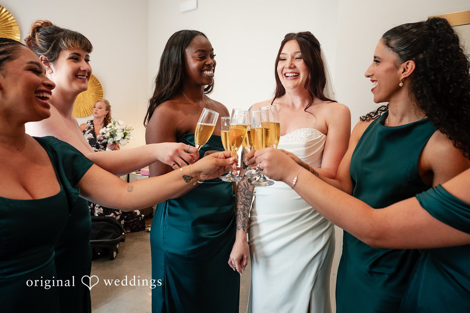 Noah + Jade A smiling bride in a white gown and four bridesmaids in deep green satin dresses toast with champagne flutes.