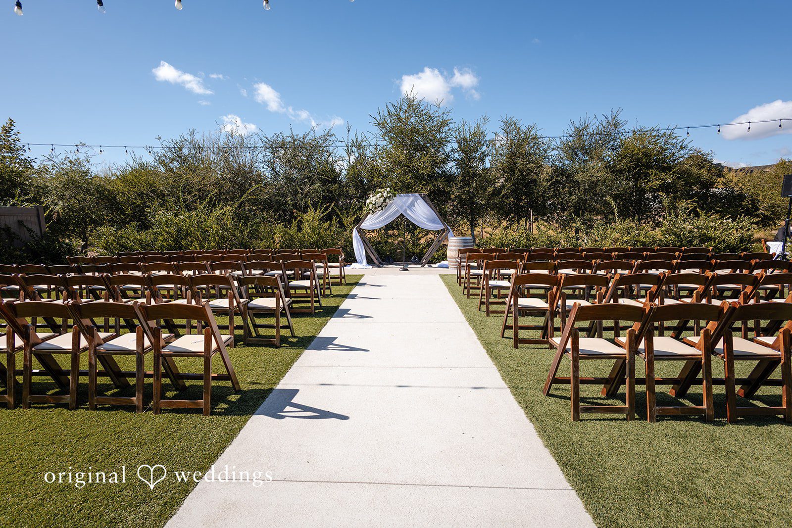 Noah + Jade Wedding ceremony setup featuring rows of wooden chairs on a lawn lining a white aisle runner that leads to a simple arch.